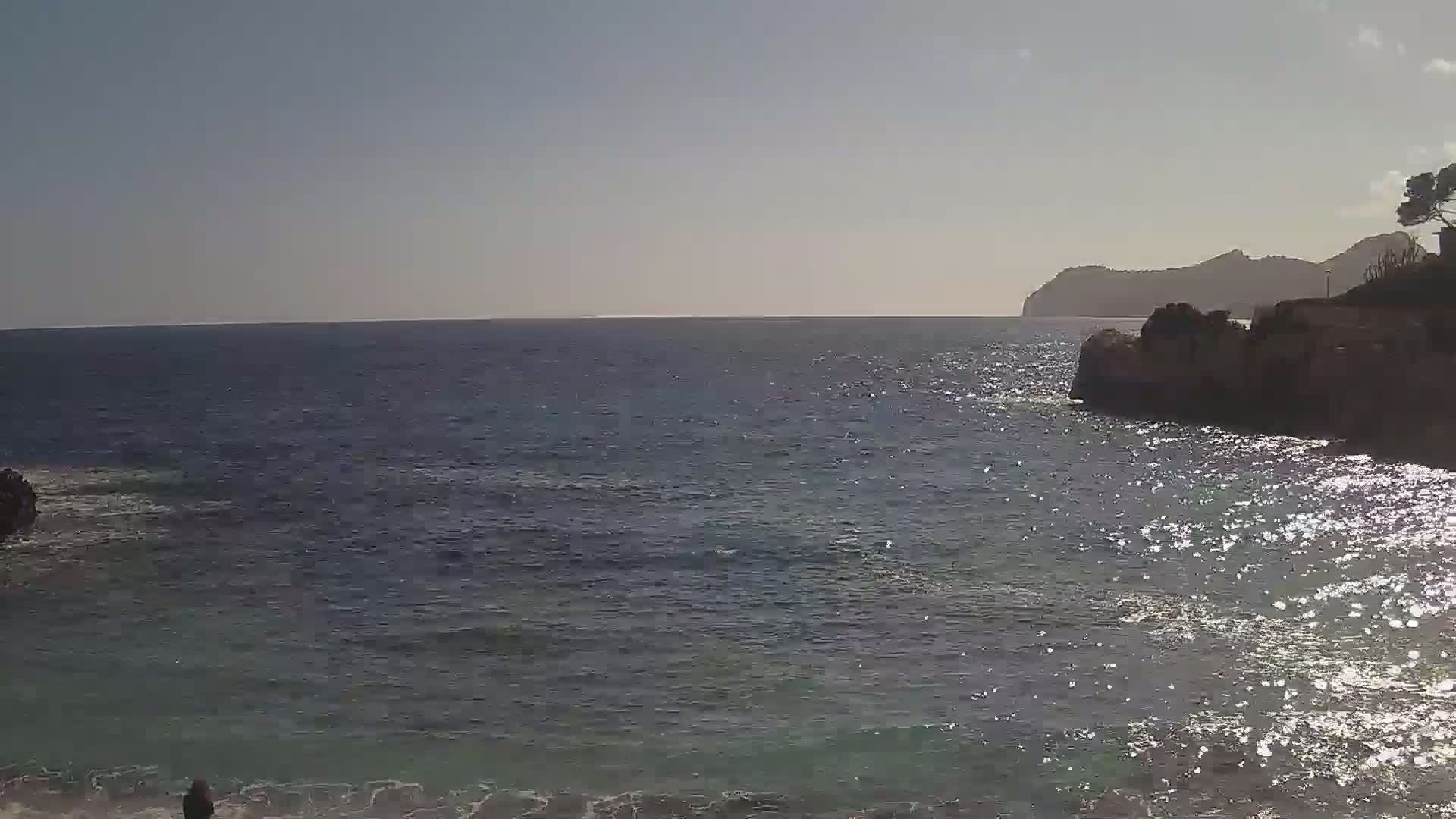 A sparkling sea laps against rocky shores under a clear sky, with a distant mountain range visible on the horizon.