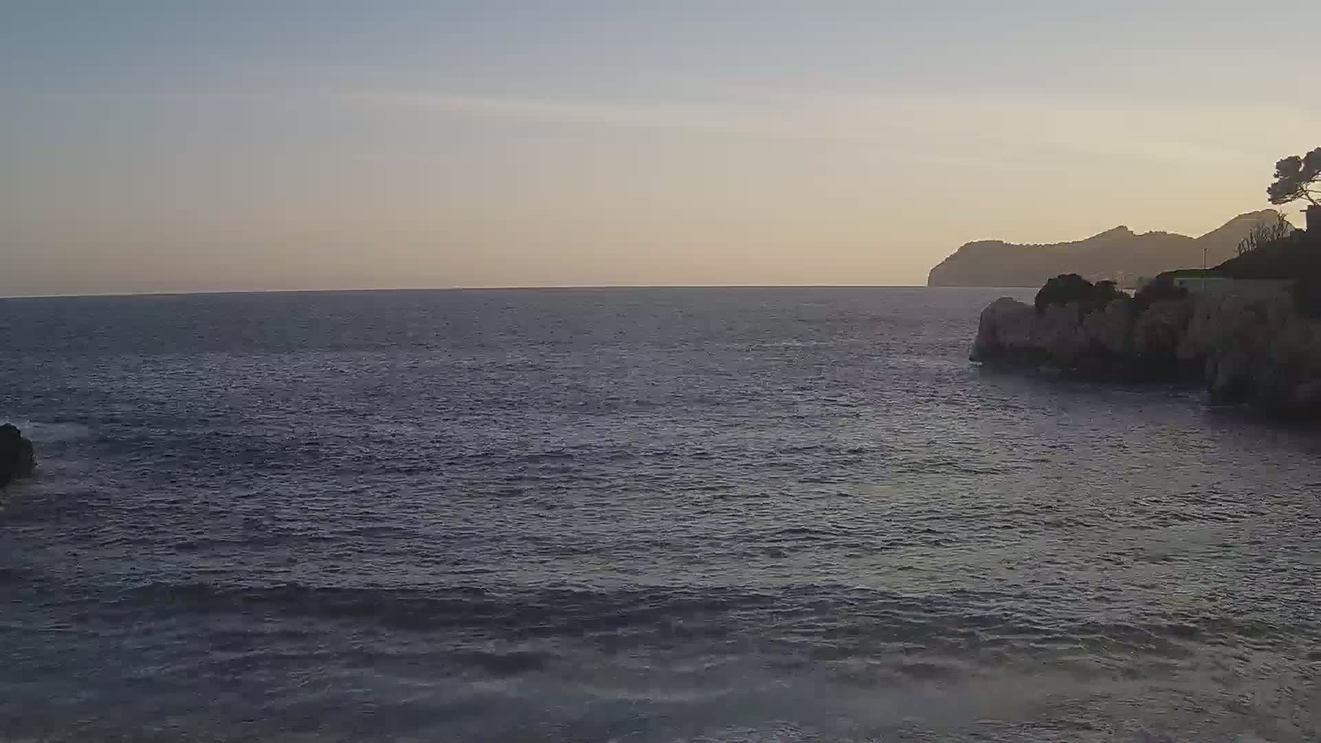 A calm sea laps against rocky shores under a clear, dusky sky, with a distant mountain range visible on the horizon.