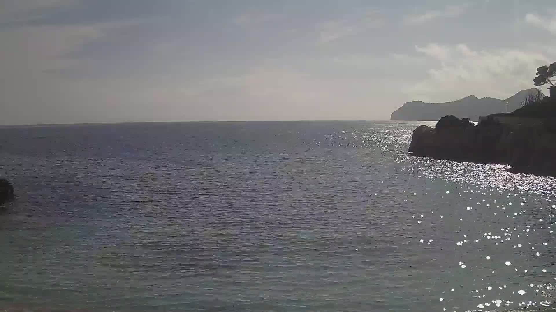 A calm sea reflecting sunlight, with a rocky shoreline and distant hills under a mostly clear sky.