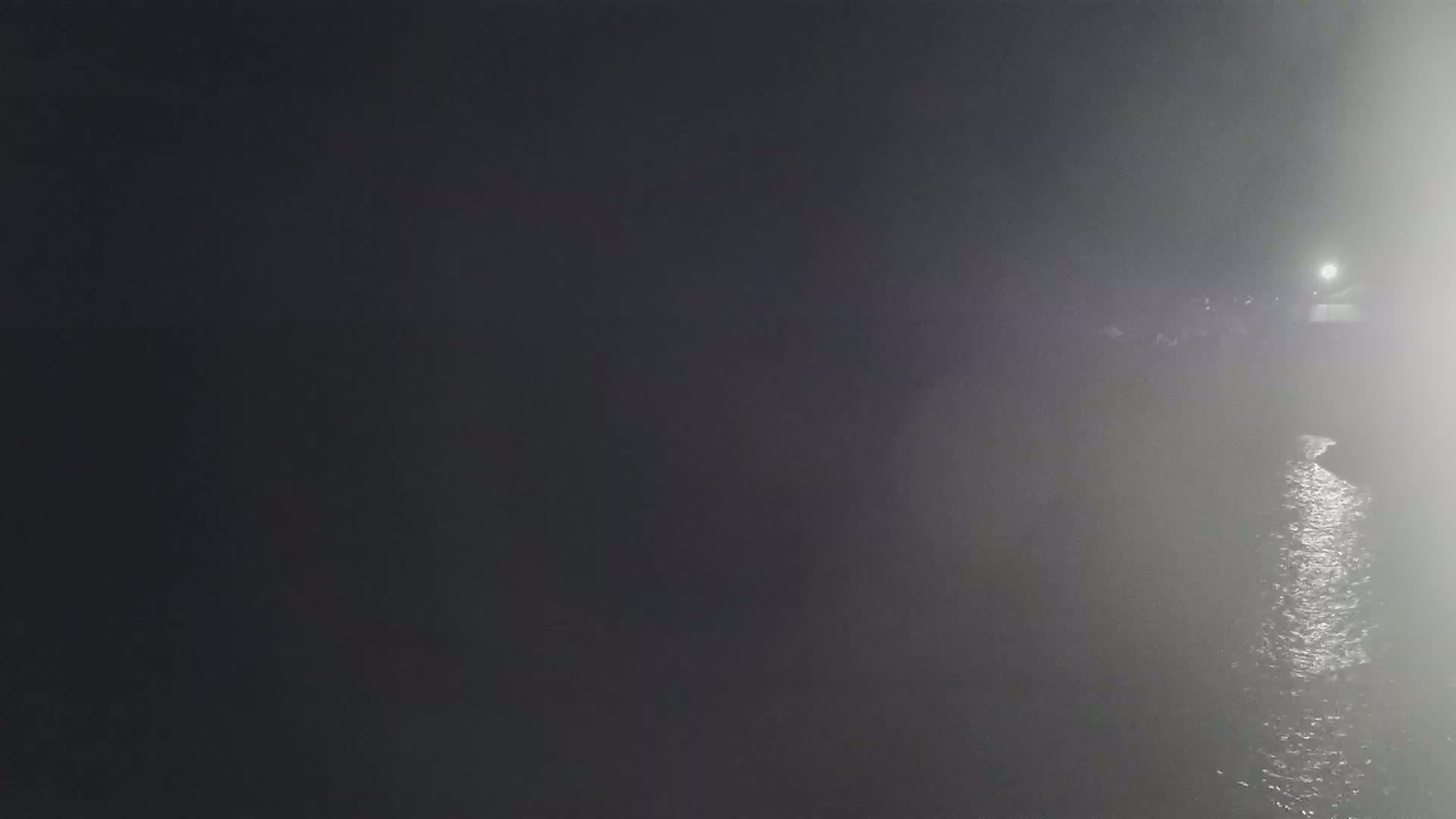 A dimly lit body of water is seen at night in foggy conditions, with a light source visible in the distance.