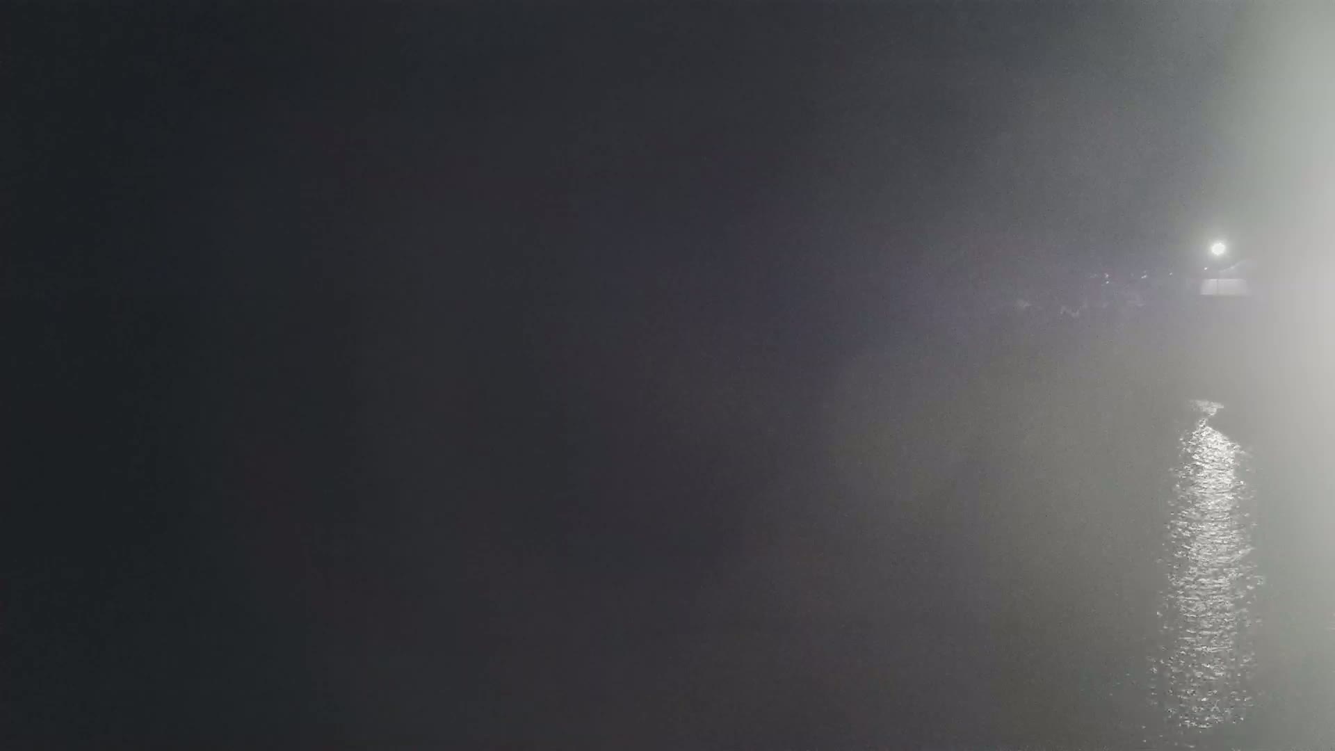 A dimly lit body of water is seen at night in foggy conditions, with a light source visible in the distance.