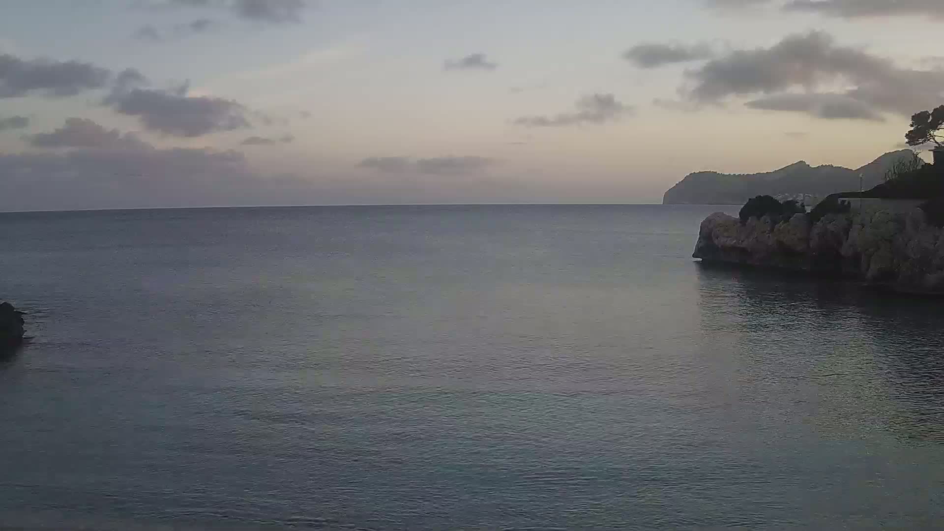 A calm, dark-blue sea under a cloudy, dusky sky meets a rocky shoreline with a distant hill in the background.