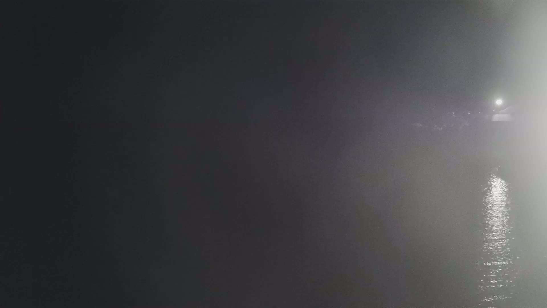 A foggy night scene shows a body of water reflecting a light source in the distance.