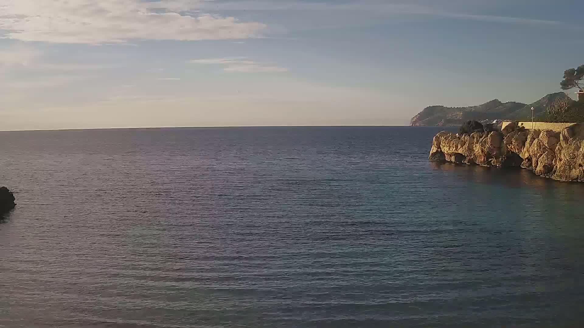 A calm sea stretches to a distant mountain range under a partly cloudy sky, with rocky cliffs and a bit of vegetation visible on the right.