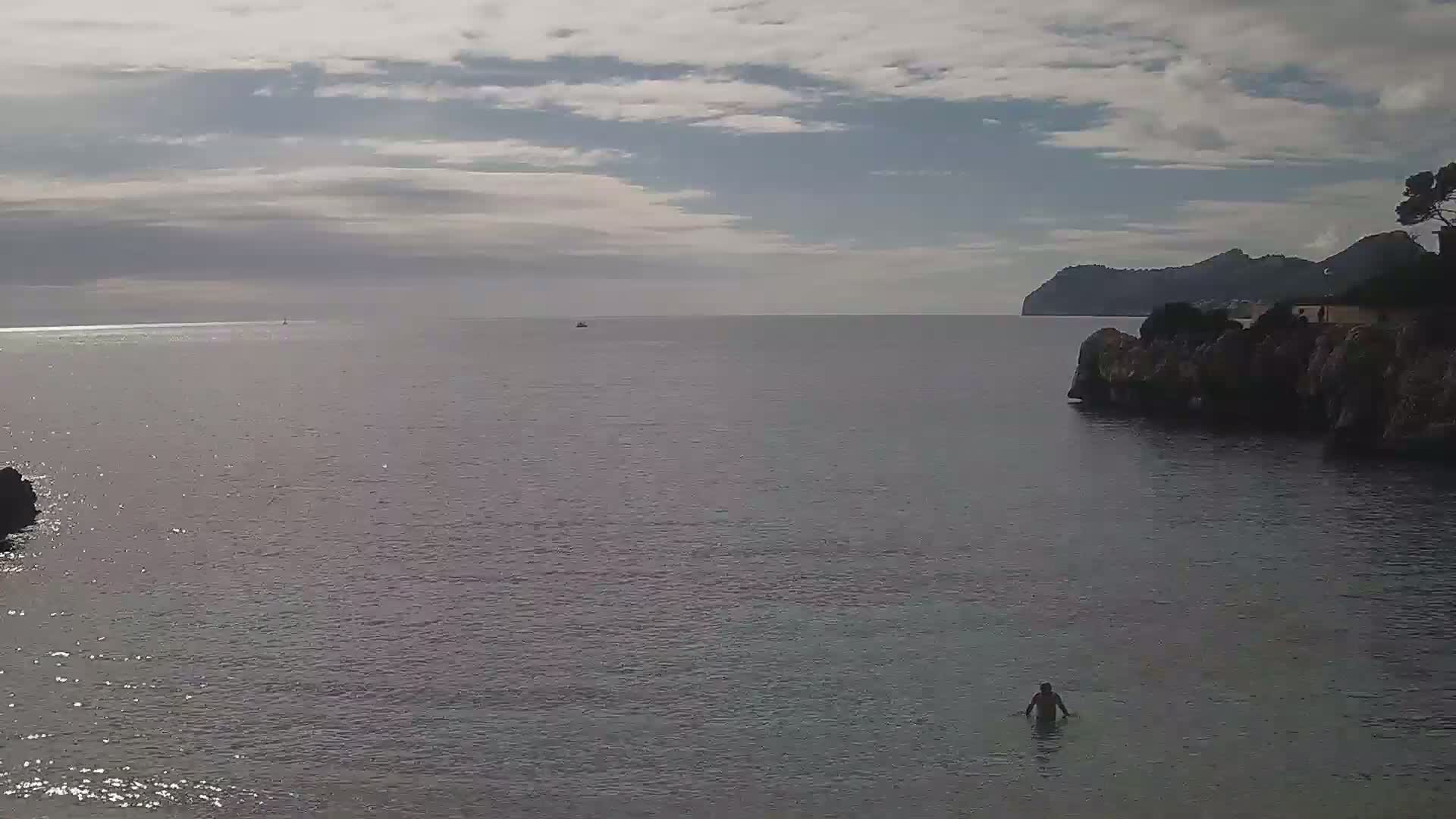 A person wades in calm, gray-blue sea water near a rocky shore, with a distant mountain range visible under a partly cloudy sky.