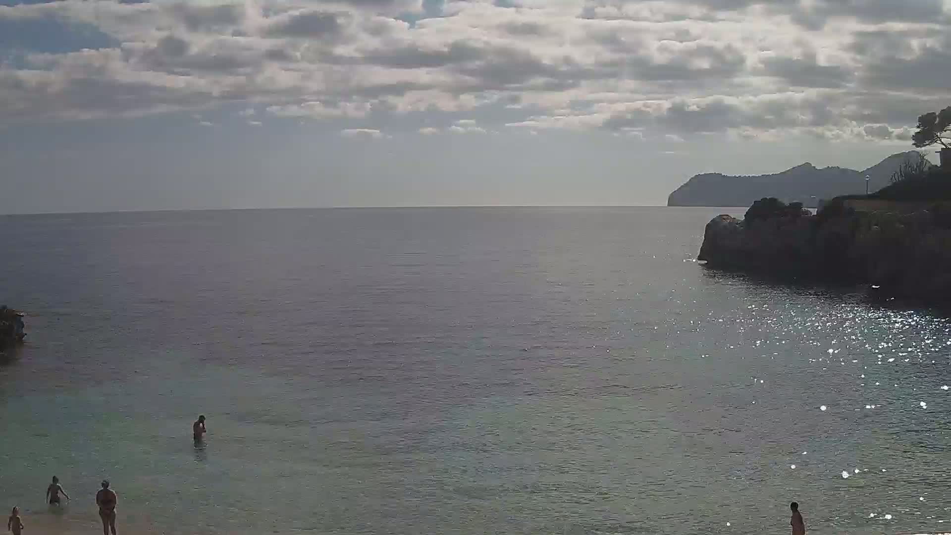 A calm, partly cloudy day at a beach with a few people swimming and wading in the shallow water, a rocky cliff on the right, and a distant mountain range visible across the sea.