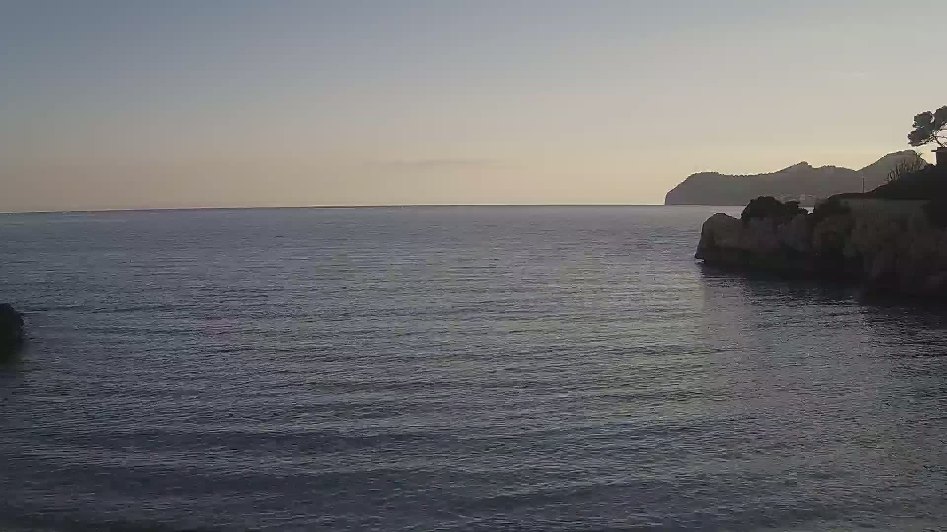 A calm, dark-blue sea laps gently against rocky shores, with a distant landmass visible under a clear, dusky sky.