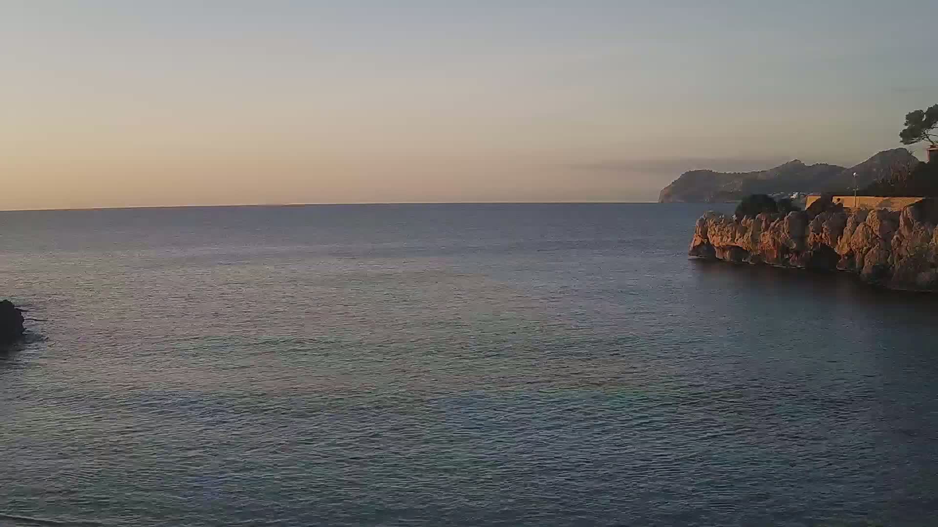 A calm sea under a pale sunset sky meets a rocky cliff top with sparse vegetation.