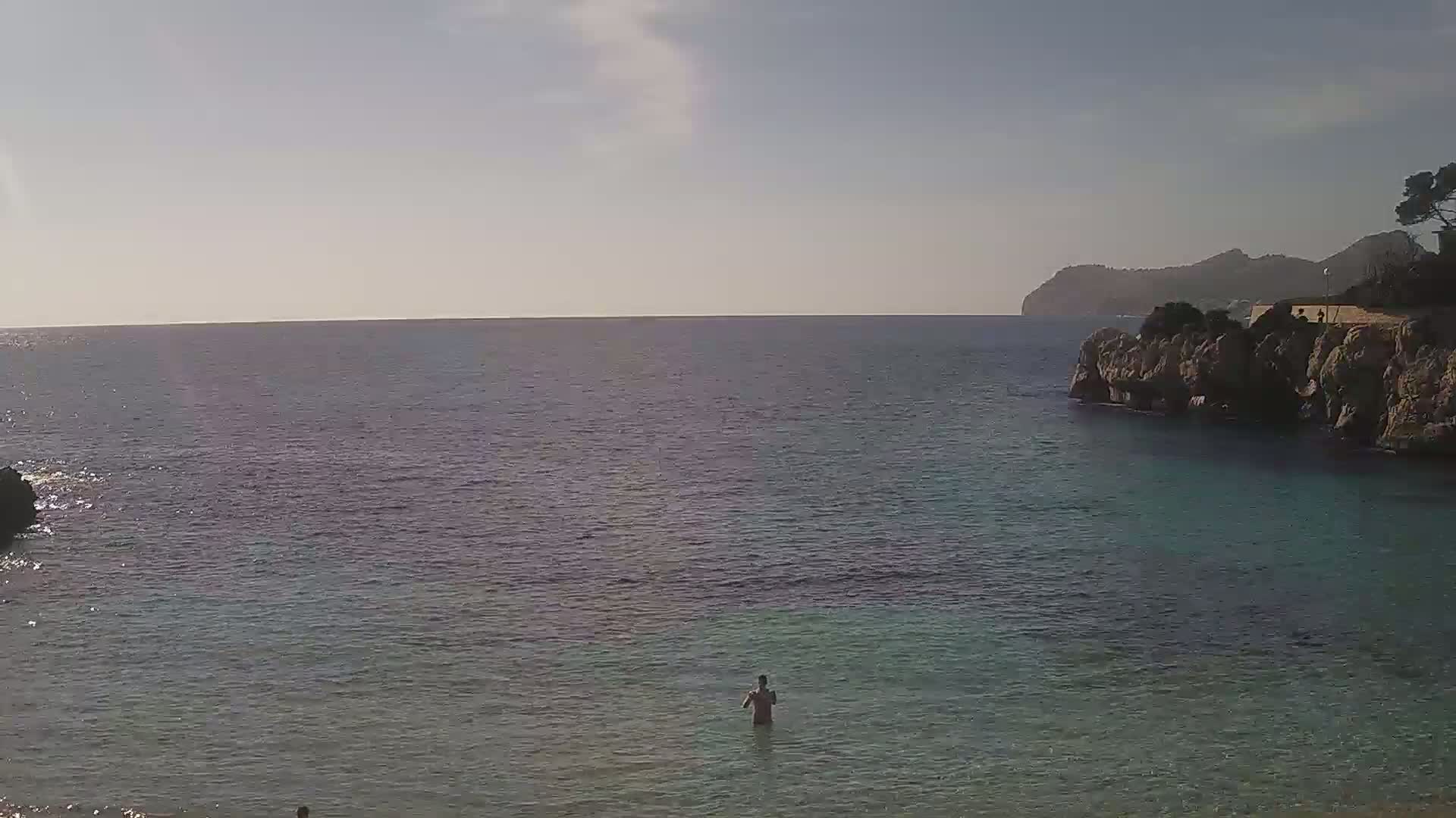 A person stands in calm, clear water near rocky shores under a mostly sunny sky.