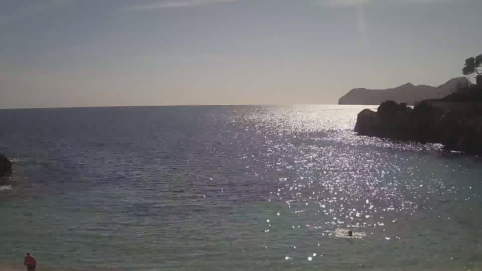 A person swims in a calm, sunlit sea near a rocky shore and a distant mountain under a clear sky.