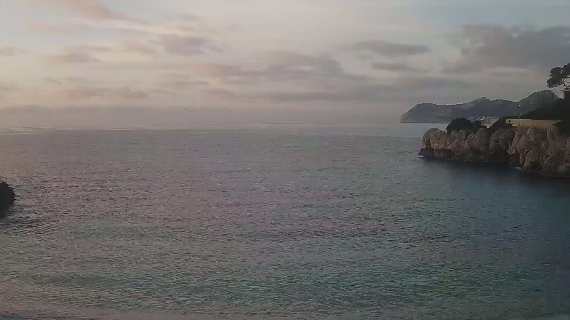 A calm sea under a cloudy, dusky sky is seen from a rocky coastline, with hills visible in the distance.