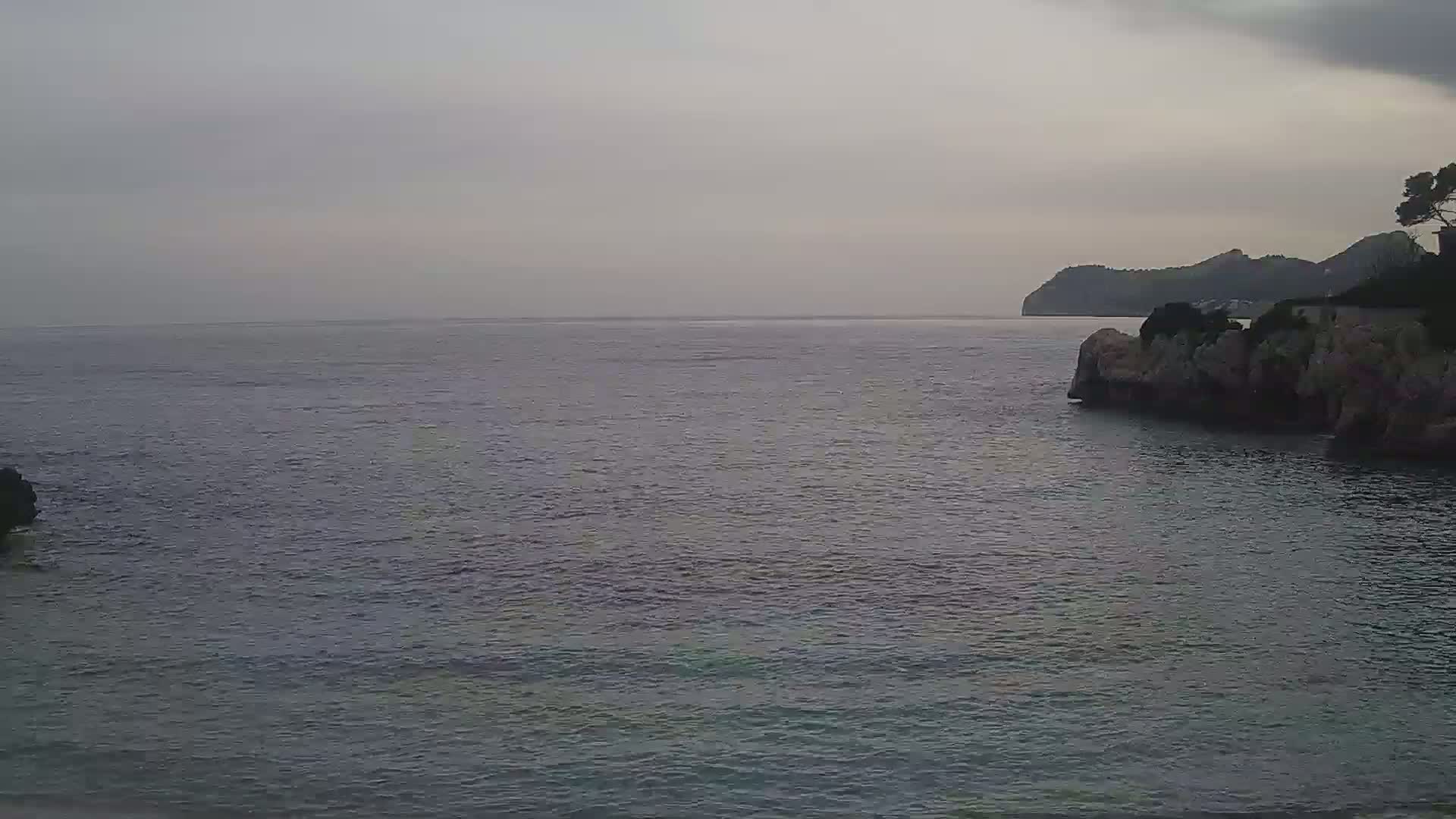 A calm, gray sea stretches to a hazy, distant shoreline under an overcast sky, with rocky outcrops visible in the foreground.
