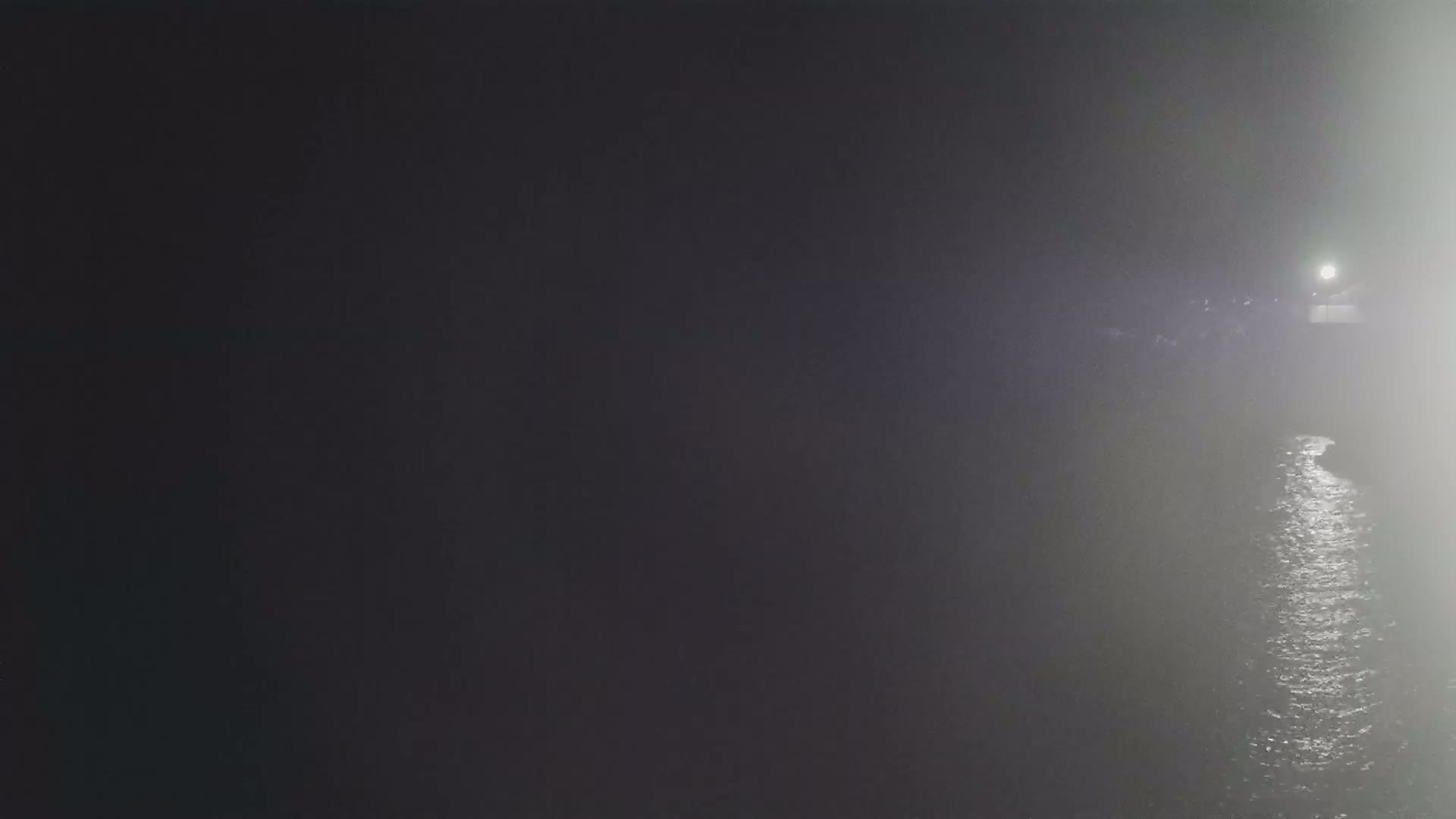A dark, foggy night scene shows a body of water reflecting a distant light source.