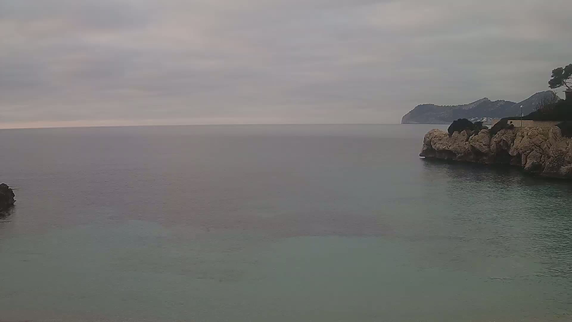A calm sea stretches to a distant mountain range under an overcast sky, with rocky outcrops at the foreground.