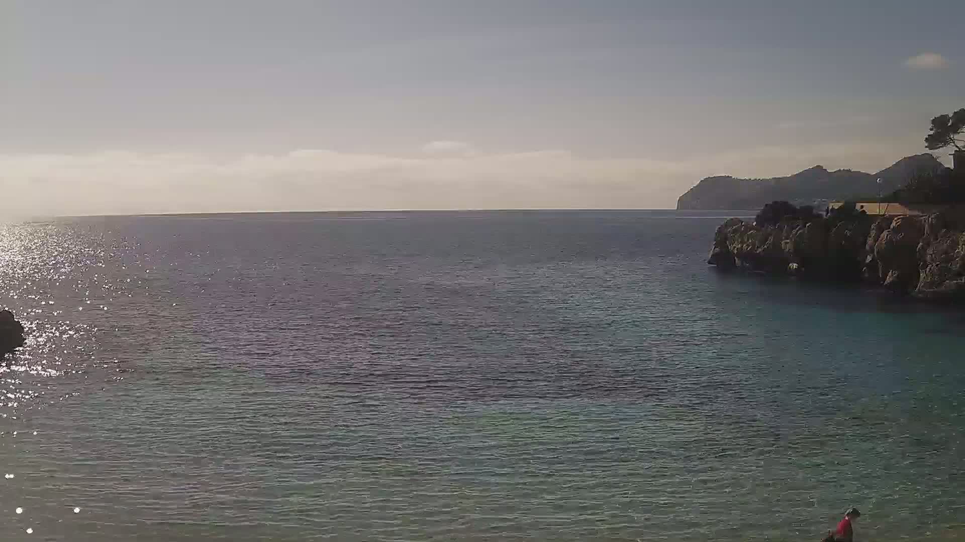 A calm, sunlit sea laps against rocky shores under a mostly clear sky, with a person visible in the lower right corner.