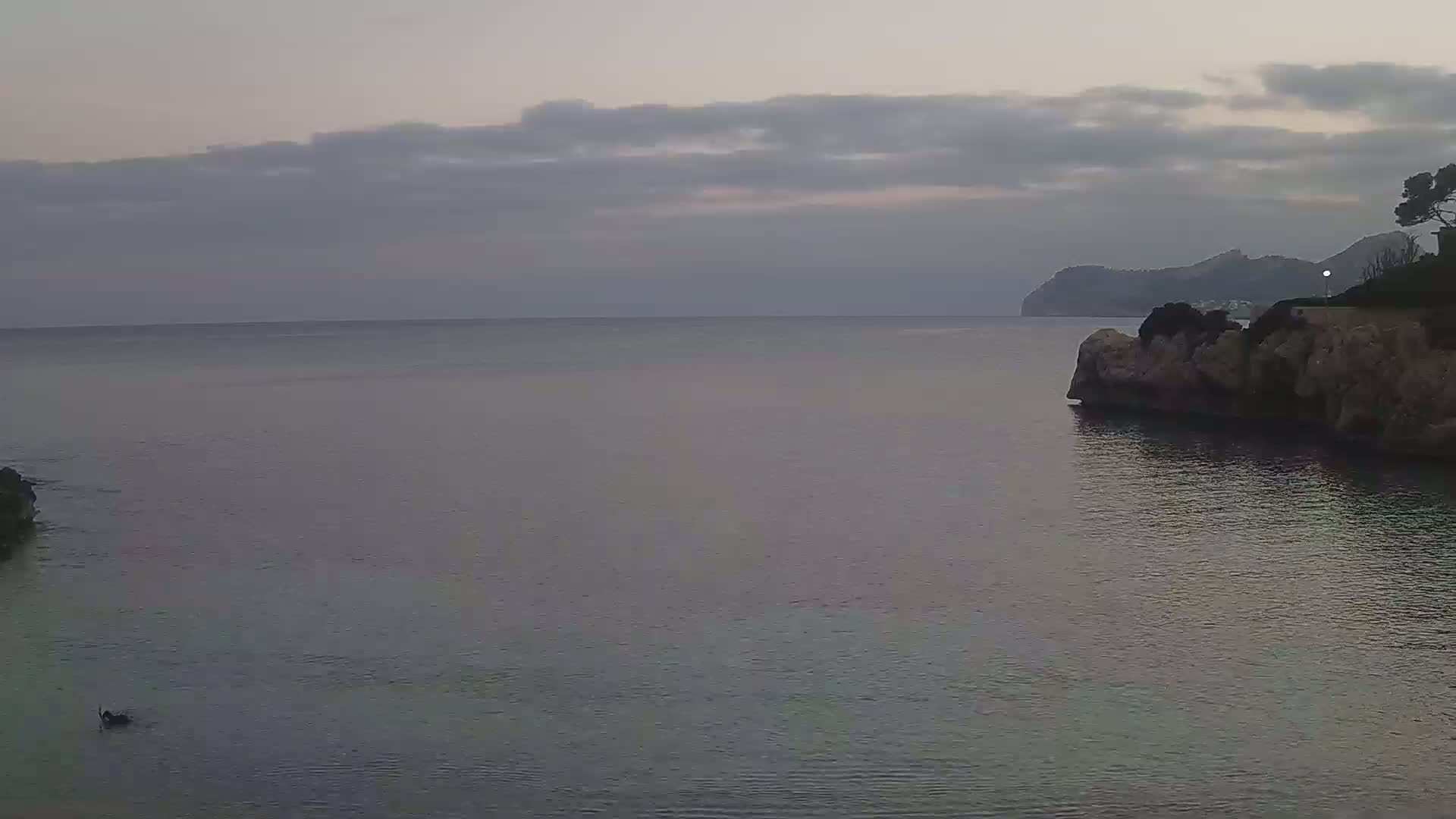 A calm, overcast sea is seen from a rocky shore, with a distant mountain range visible under a cloudy sky.