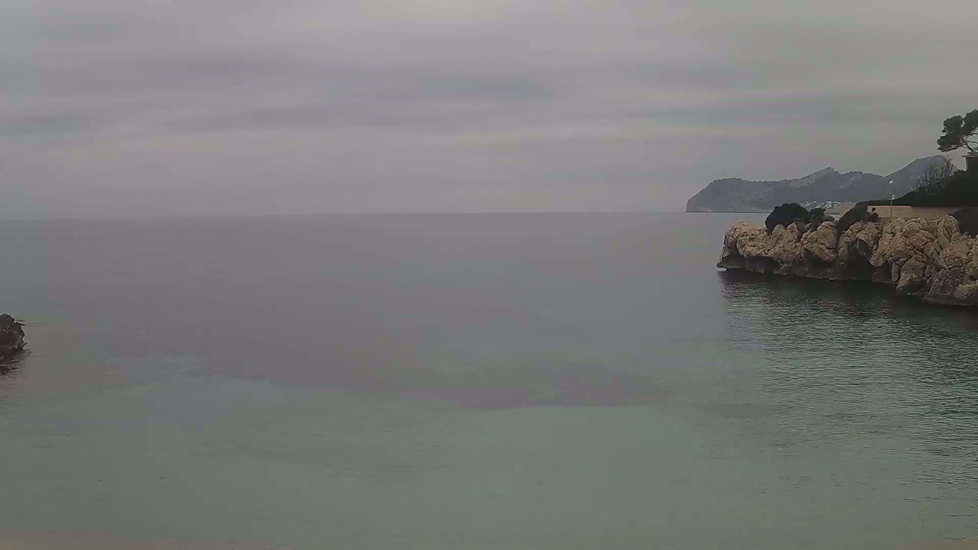 A calm, overcast day shows a rocky shoreline and teal water with a distant mountain range visible under a grey sky.