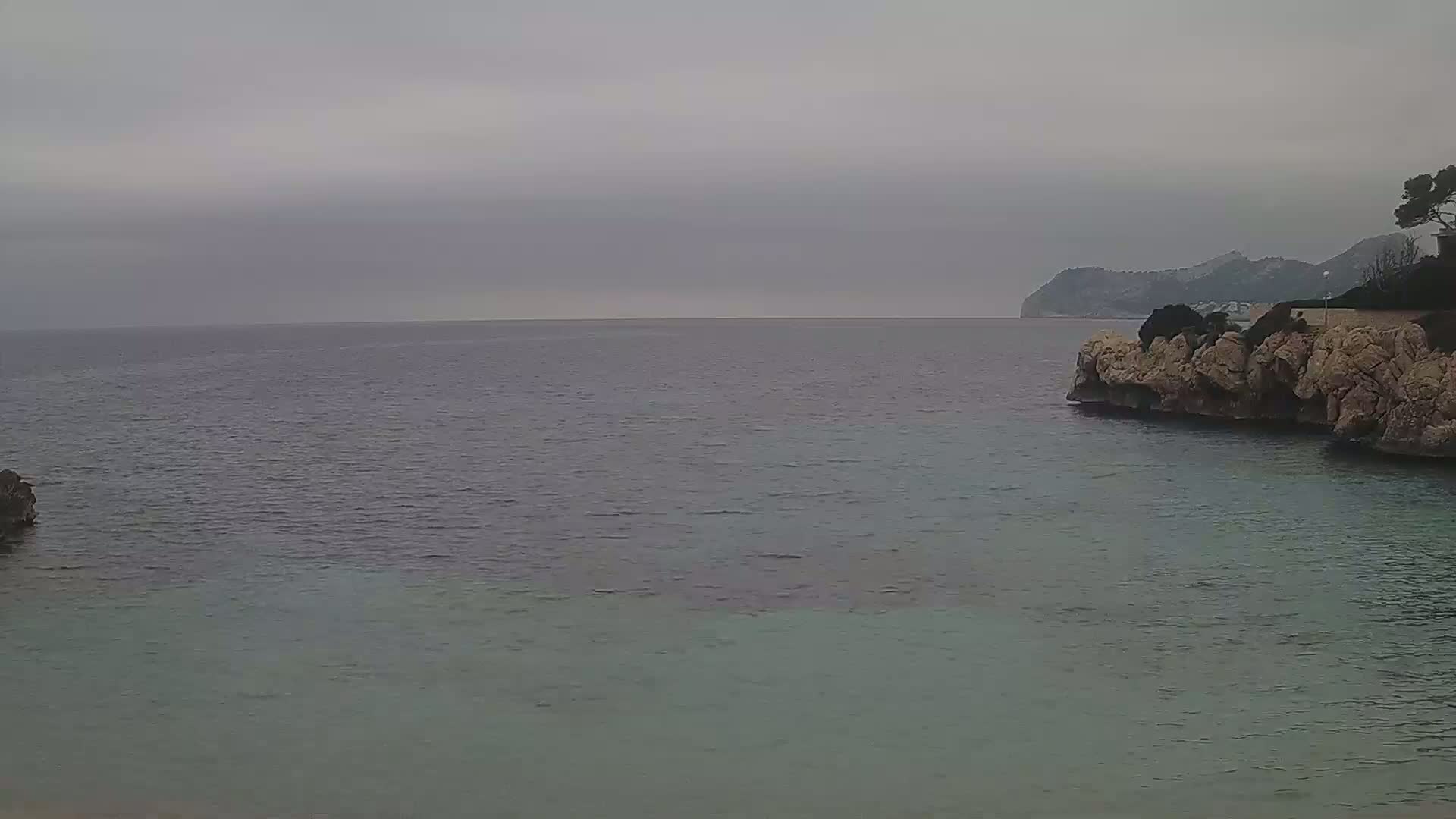A calm, overcast day shows a rocky shoreline with a body of water extending to a distant mountain range.