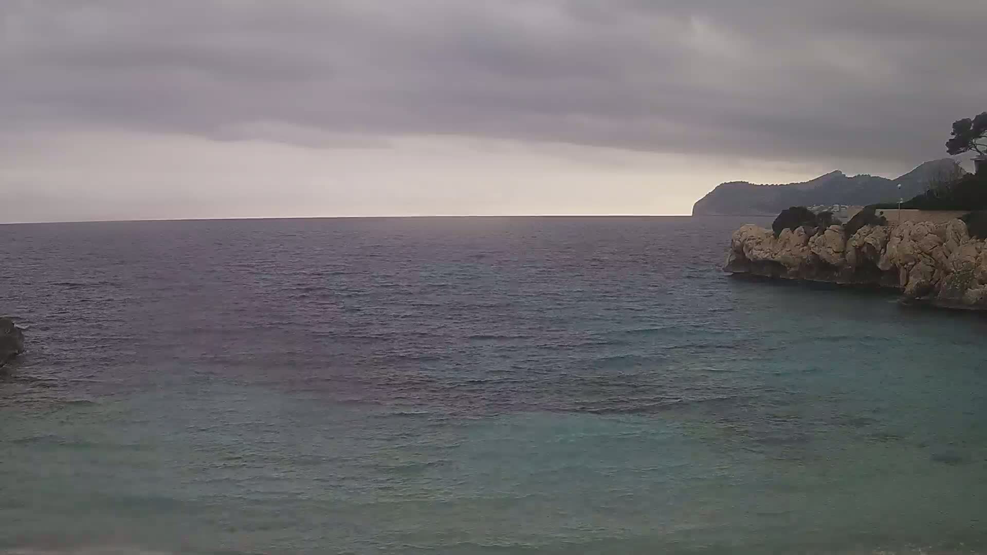 A rocky coast meets a calm, dark-blue sea under a cloudy, overcast sky.