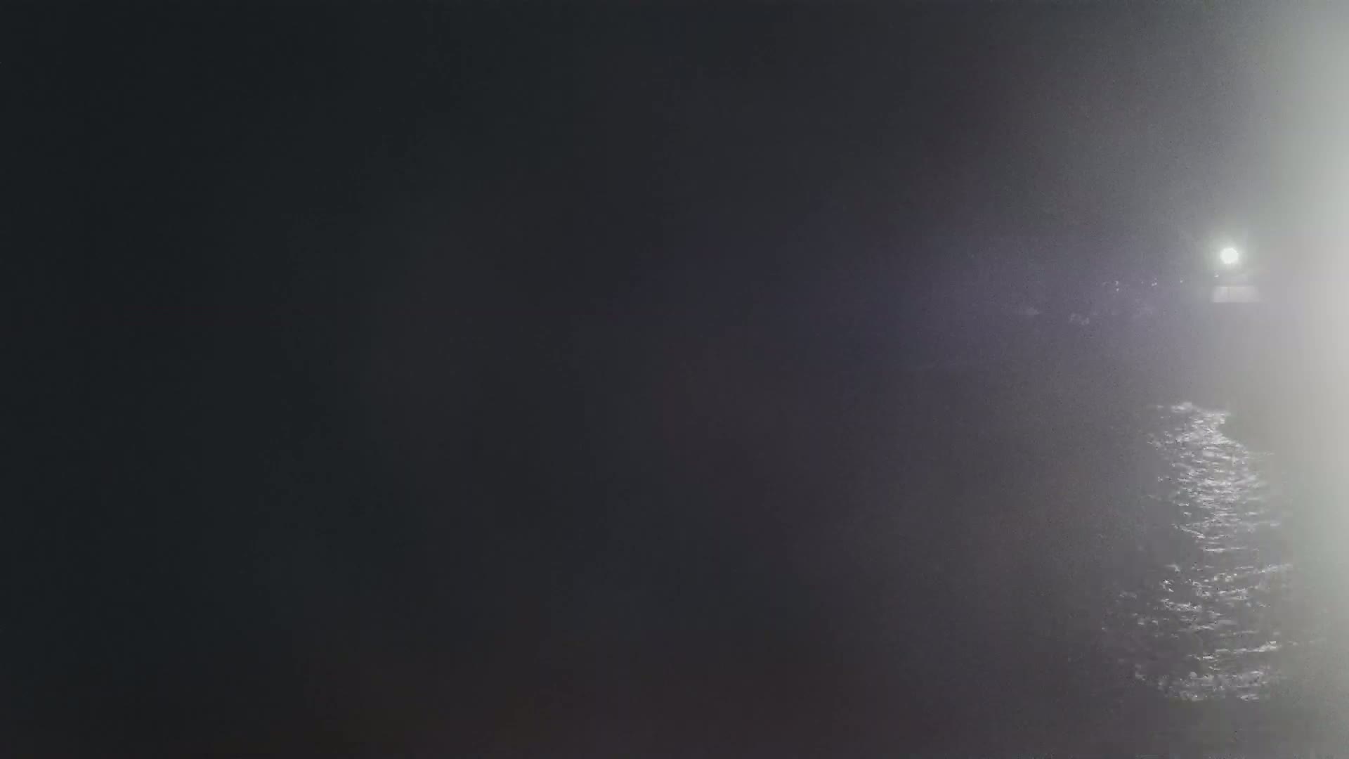 A bright light is visible in the distance through a thick fog at night over a dark body of water.