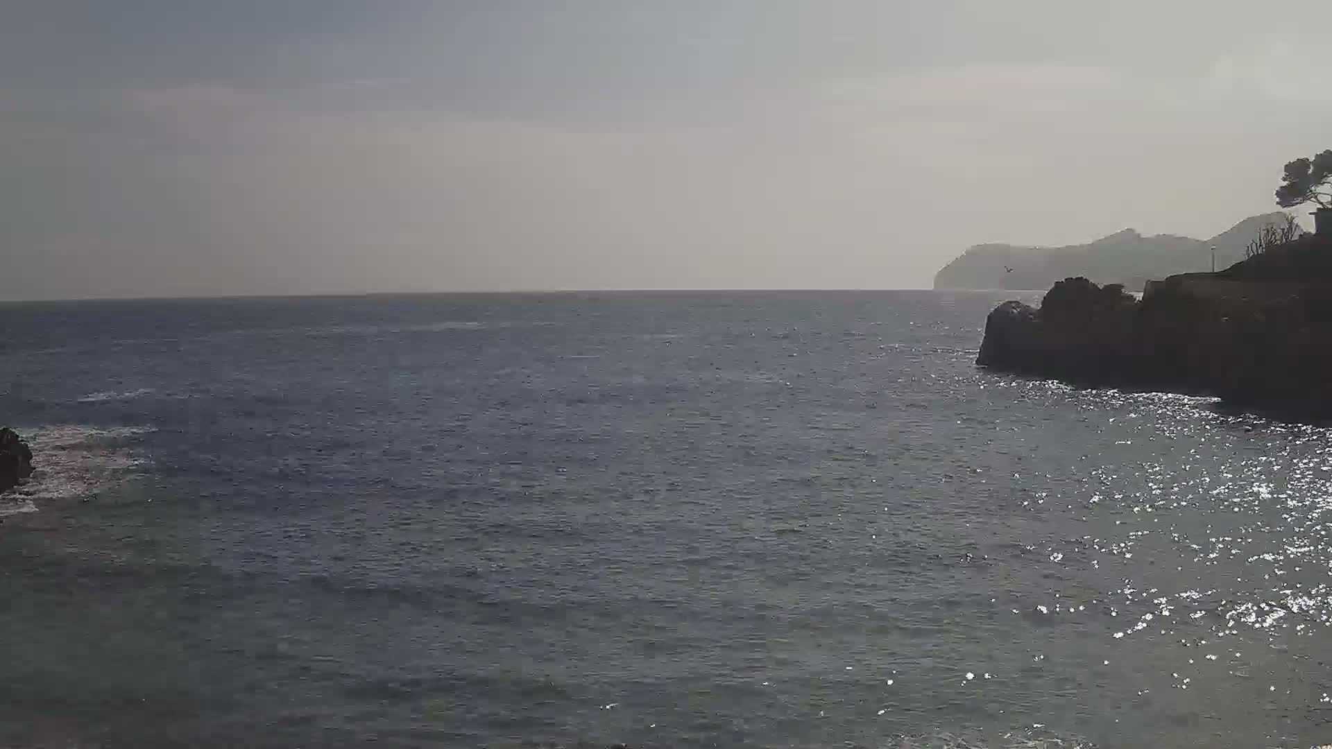 A calm, sunlit sea laps against dark rocky cliffs, with a distant hazy landmass visible on the horizon.