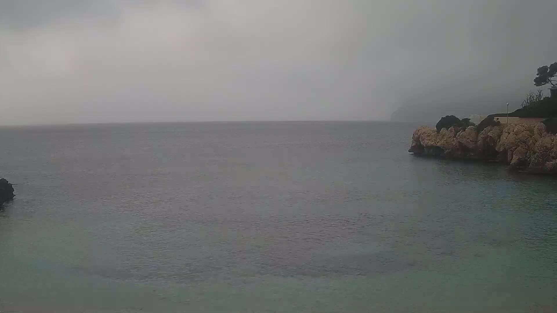 A calm, grey sea is seen from a rocky cove on an overcast day.