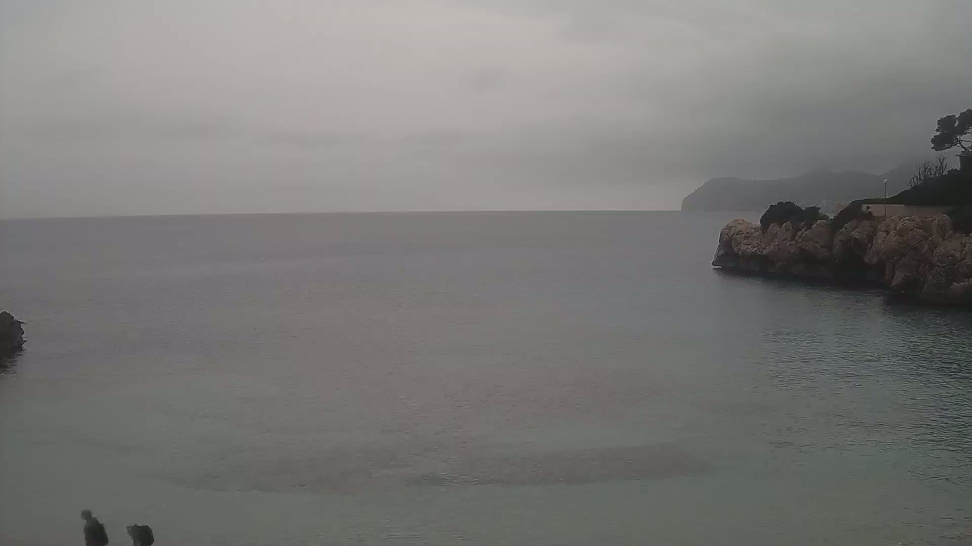 A calm, grey sea stretches to a hazy, distant shoreline under an overcast sky, with rocky points of land on either side of the foreground.