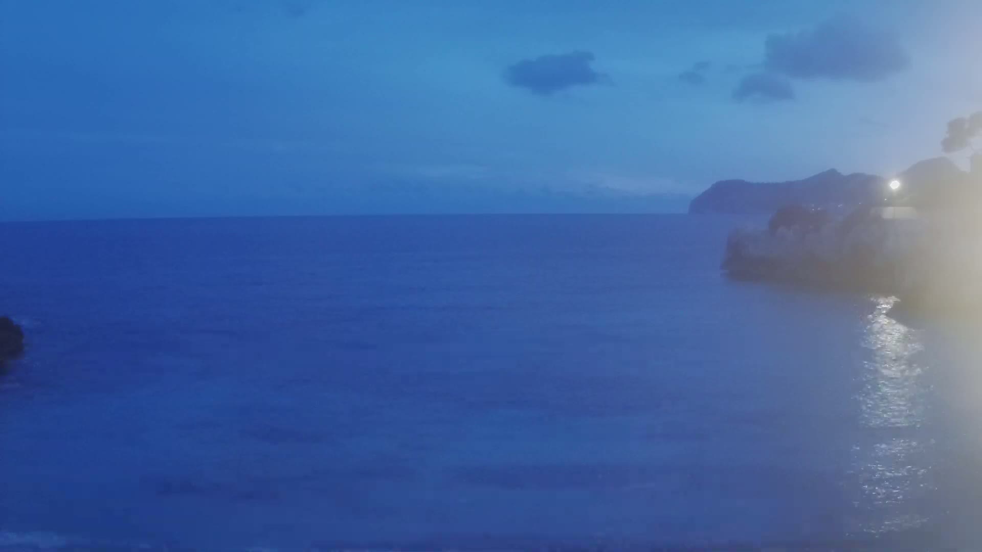 A dark blue sea at dusk under a mostly clear sky meets a rocky shoreline with a distant, silhouetted hill.