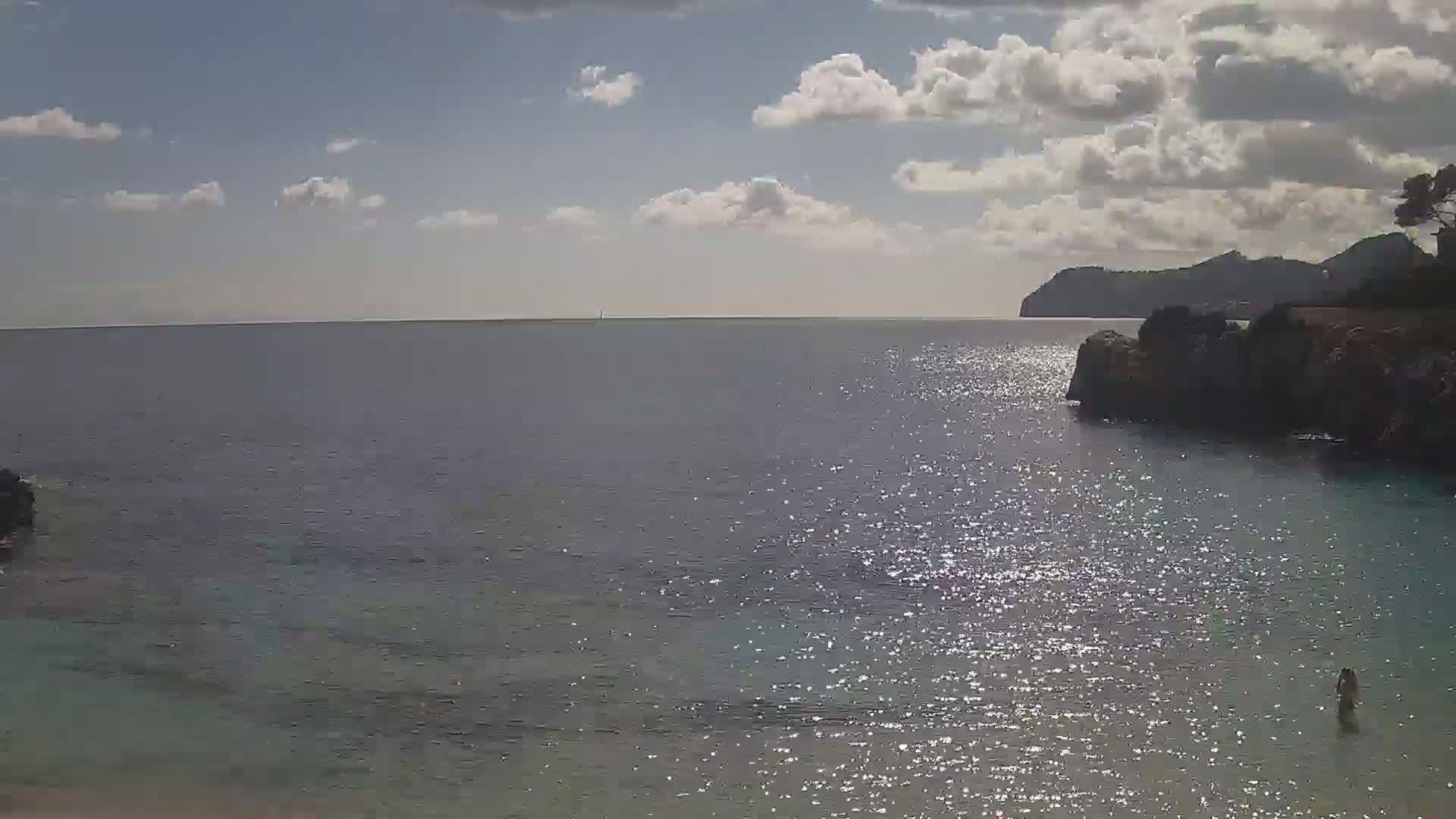 A partly cloudy day shows a calm, sparkling sea with a person wading in the shallows near a rocky shoreline and a distant mountain.