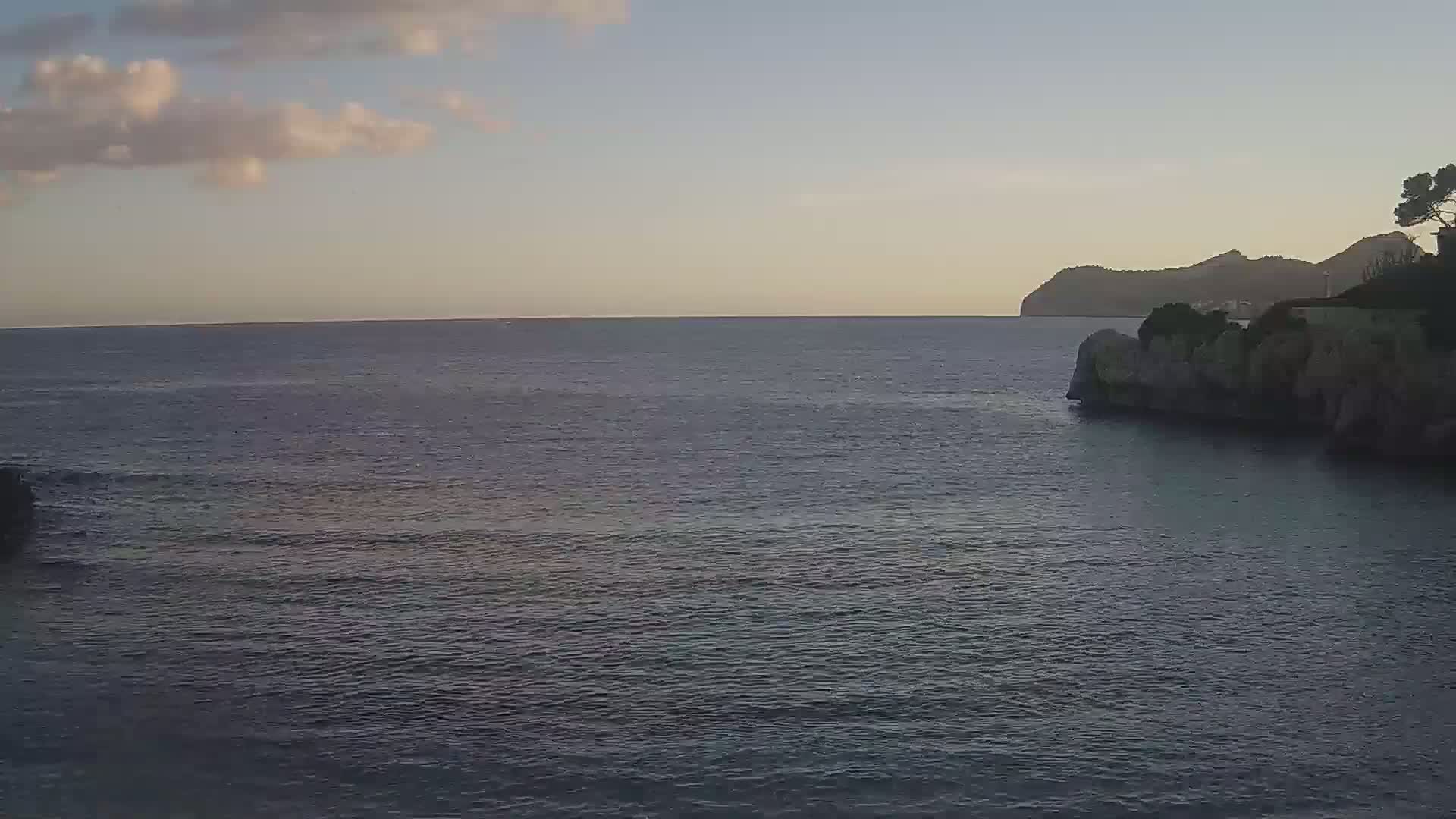 A calm, dark-blue sea laps gently against rocky shores under a partly cloudy, dusky sky, with a distant mountain visible on the horizon.