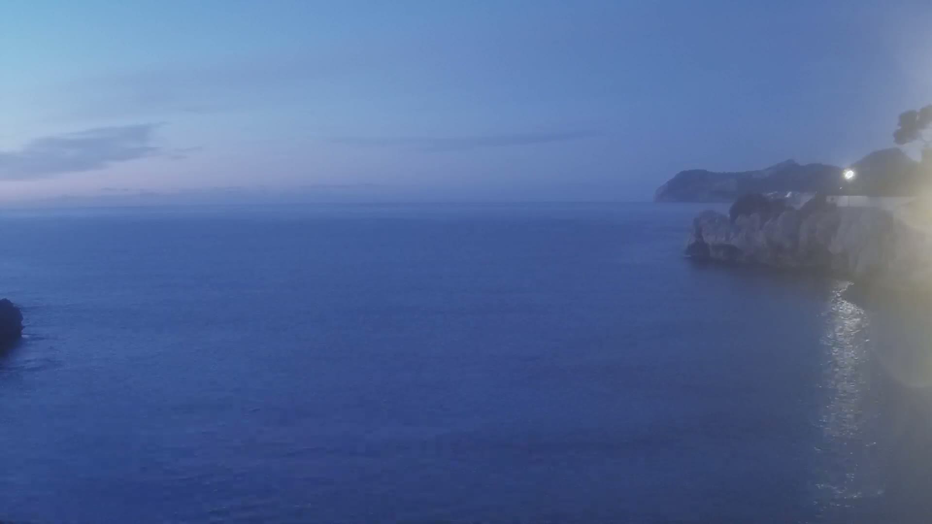 A calm, dark blue sea at twilight laps against rocky shores under a mostly clear sky.