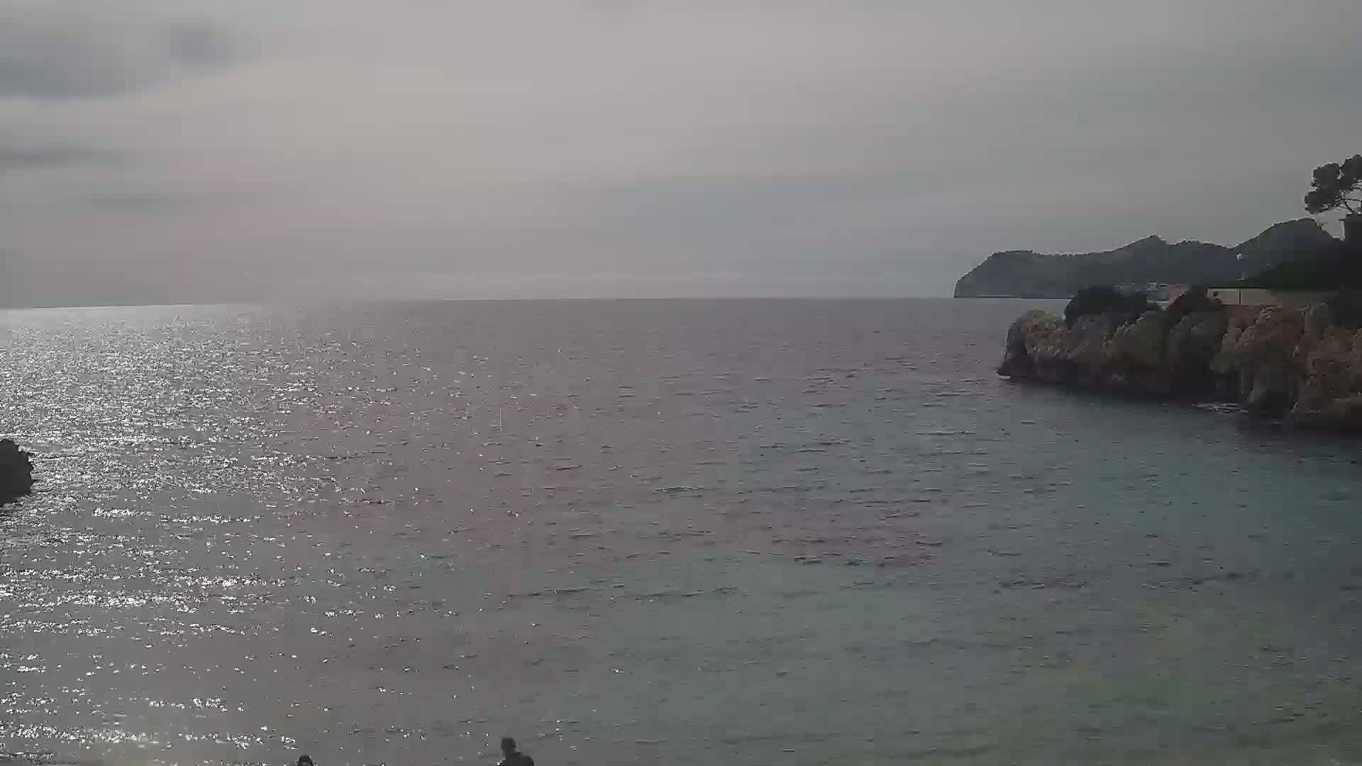 A calm, gray sea laps against a rocky shore, with a distant hill visible under an overcast sky.