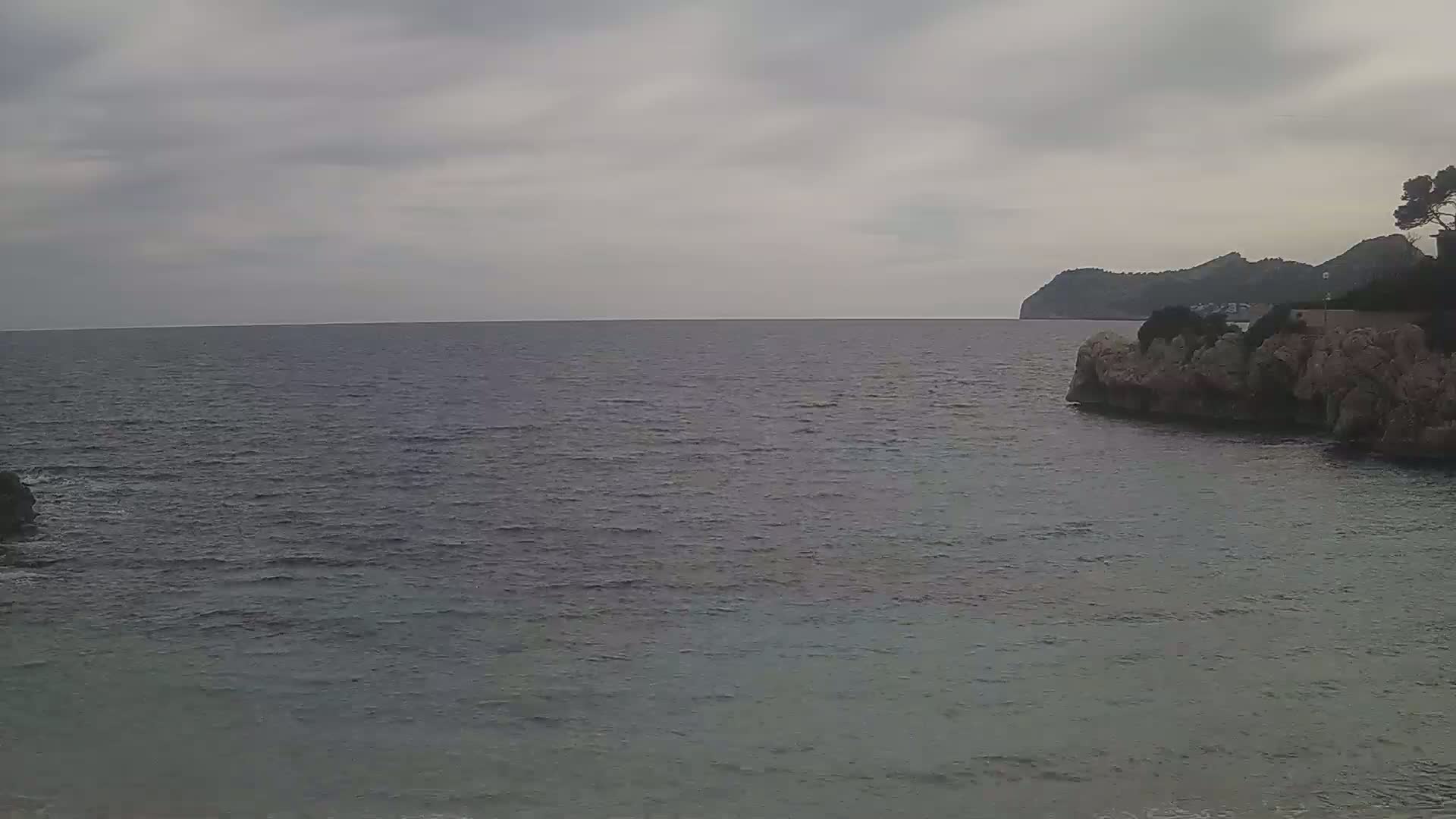 A calm sea stretches to a distant, tree-topped hill under an overcast sky.