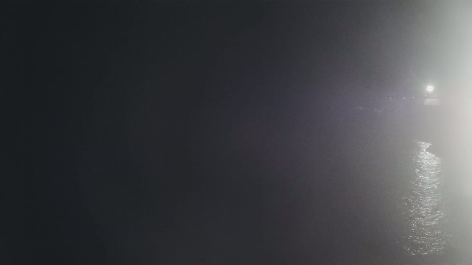 A dimly lit body of water is seen at night in foggy conditions, with a light source visible in the distance.