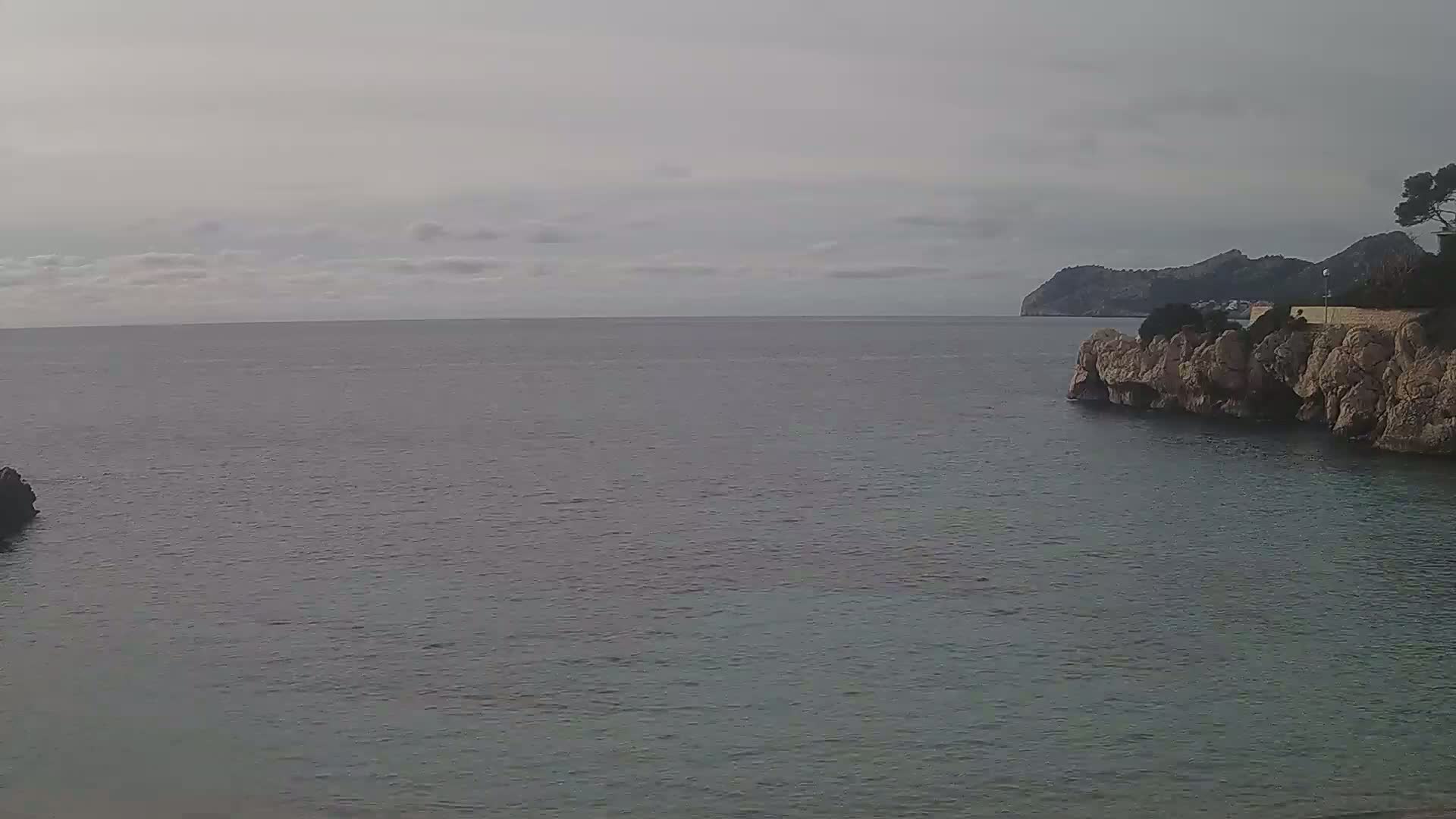 A calm sea under an overcast sky is partially enclosed by rocky shores, with a distant mountain visible on the horizon.