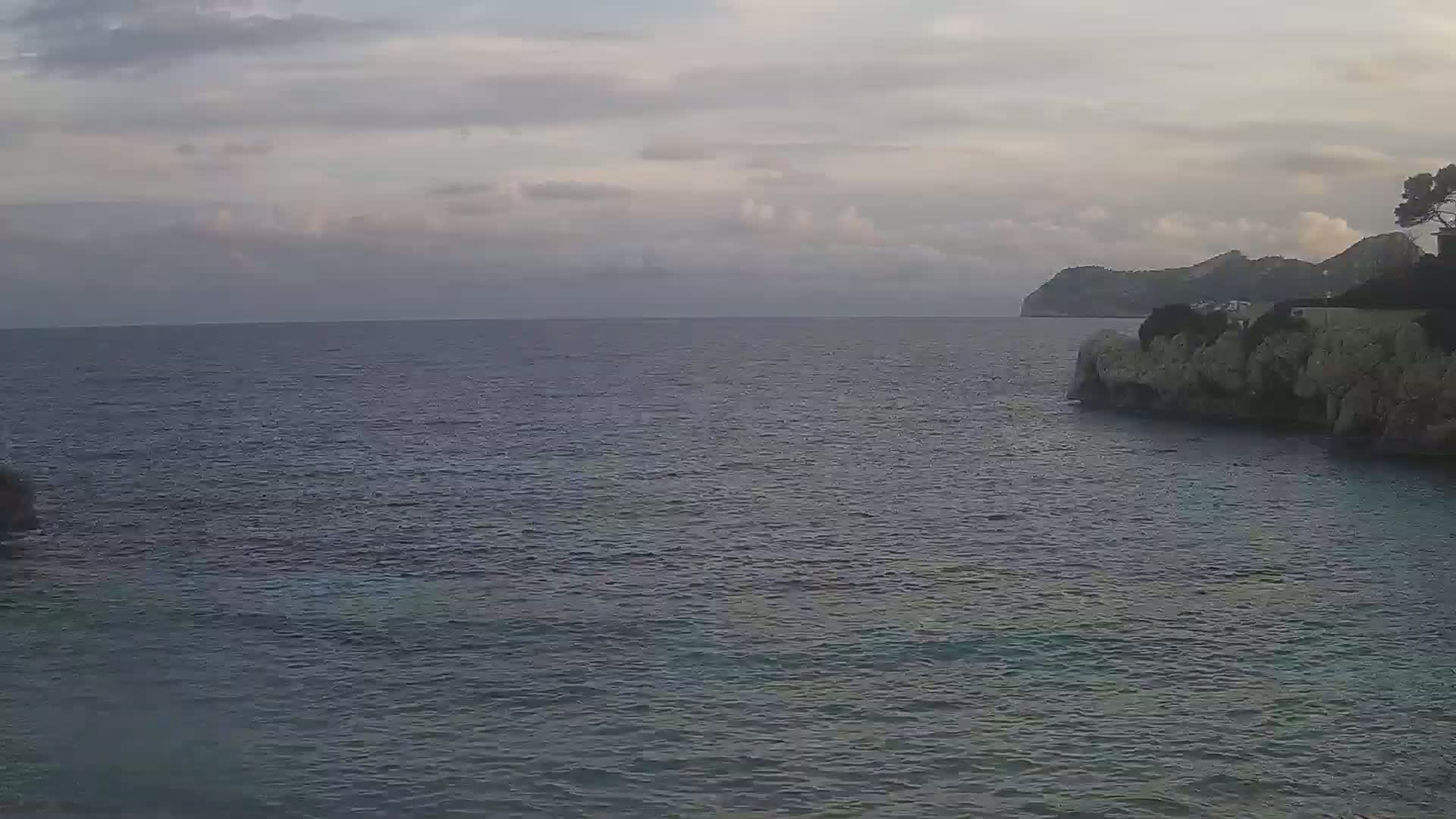A calm, dark-blue sea stretches to a distant, rocky shoreline under a cloudy sky.