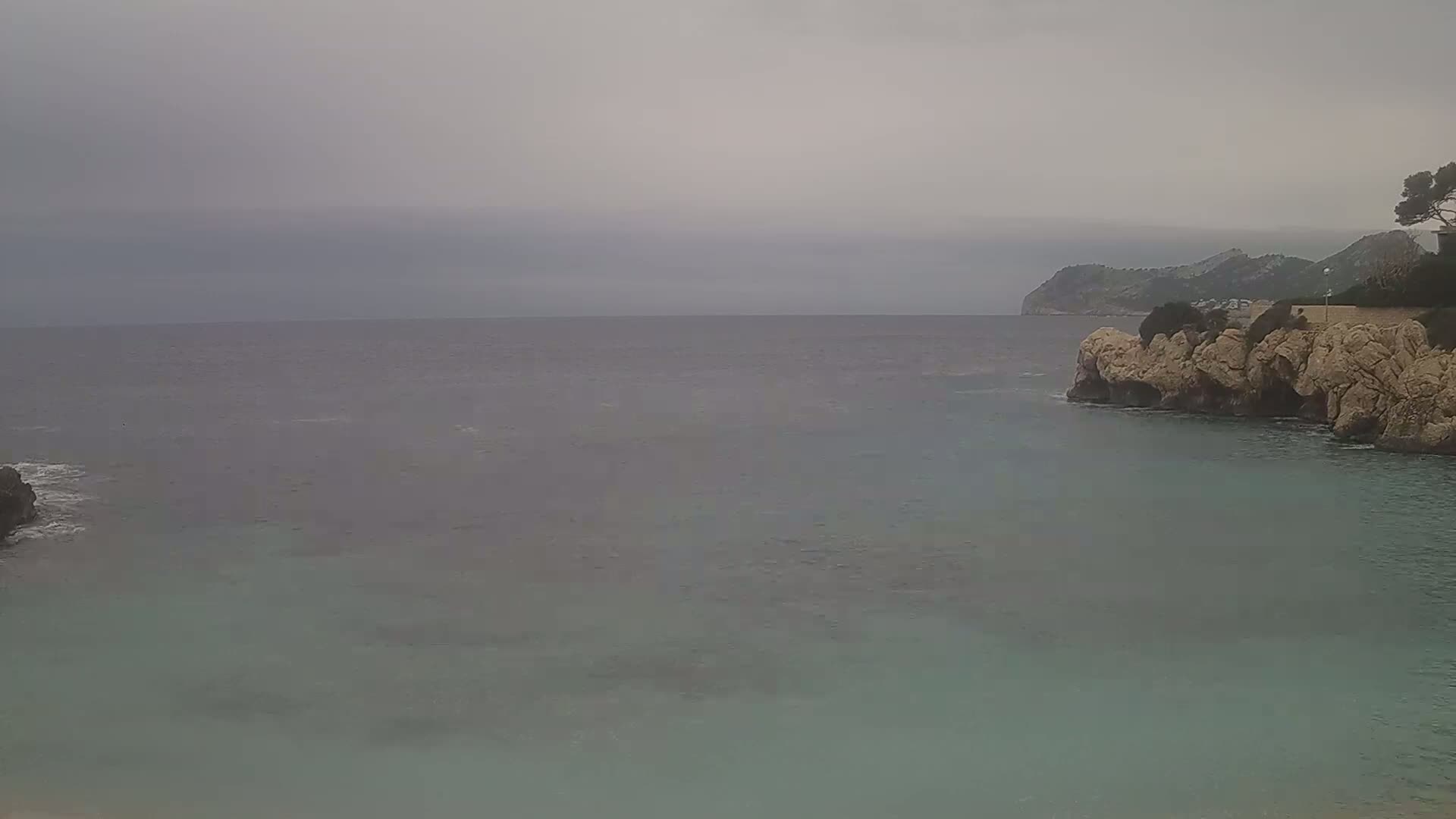 A calm, overcast day shows a rocky cove with teal water meeting a gray sea under a pale sky.