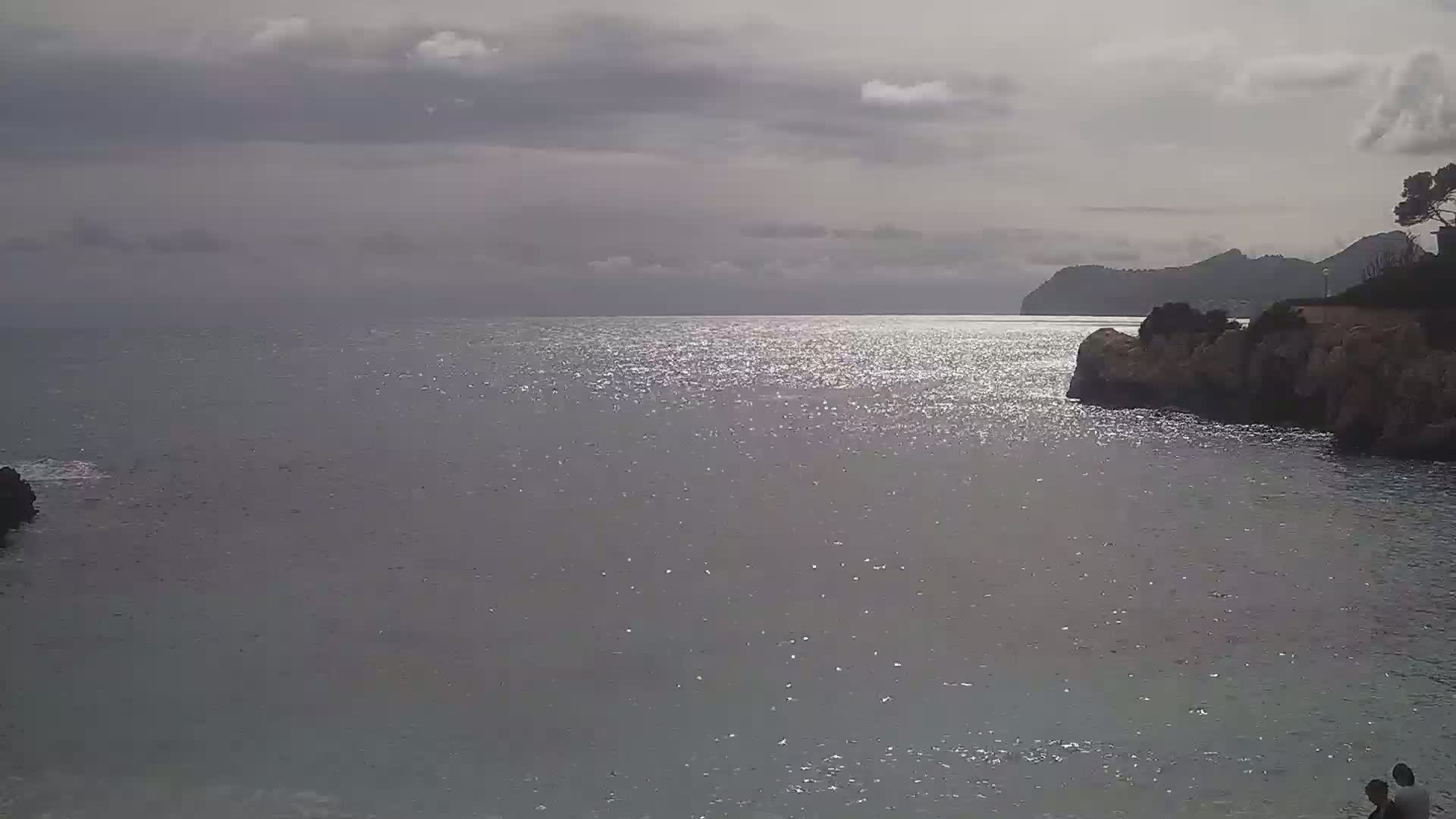 A calm, sunlit sea stretches to a distant, mountainous shoreline under a mostly cloudy sky.