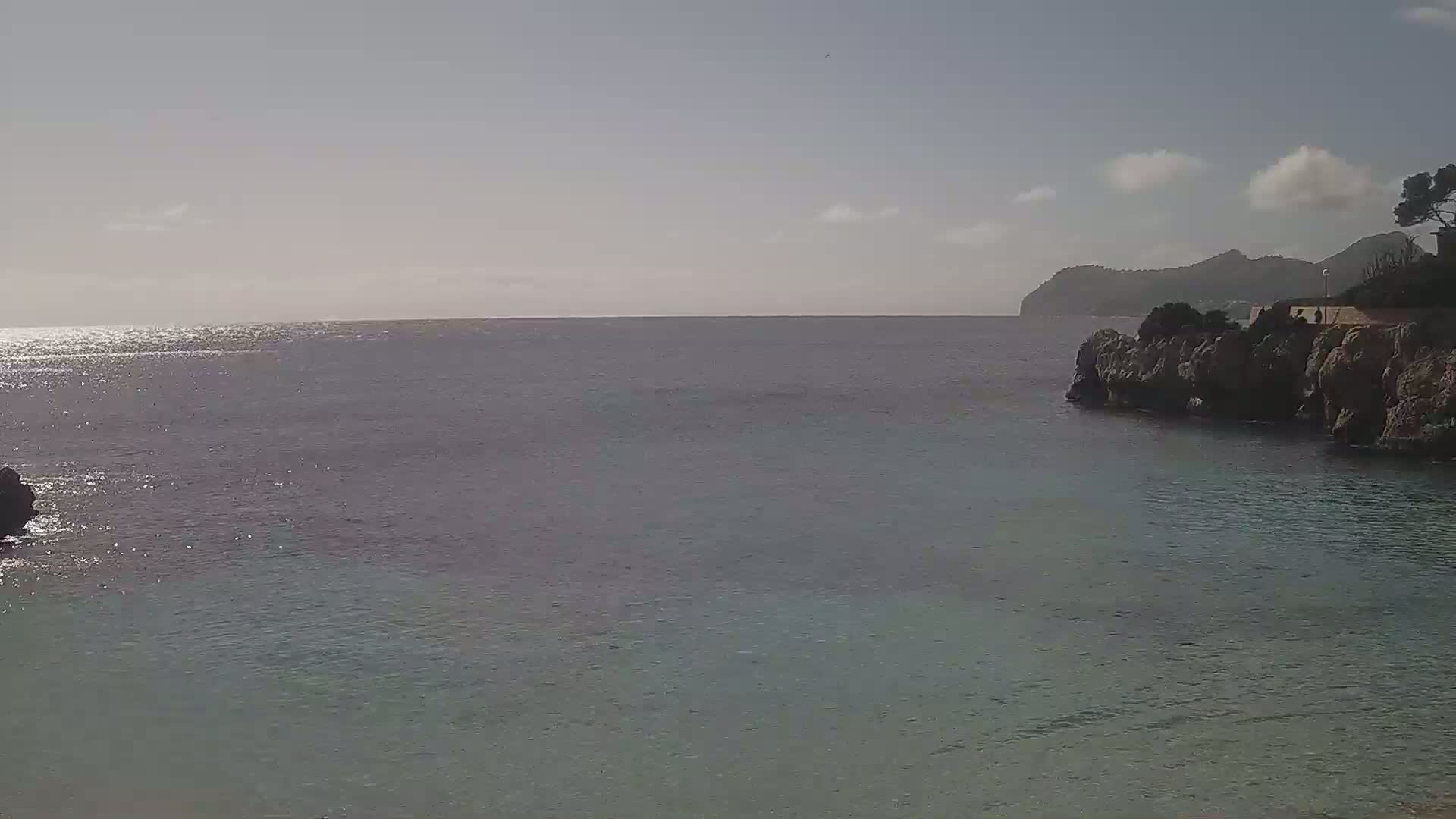 A calm, sunlit sea laps against rocky shores, with distant mountains visible under a partly cloudy sky.