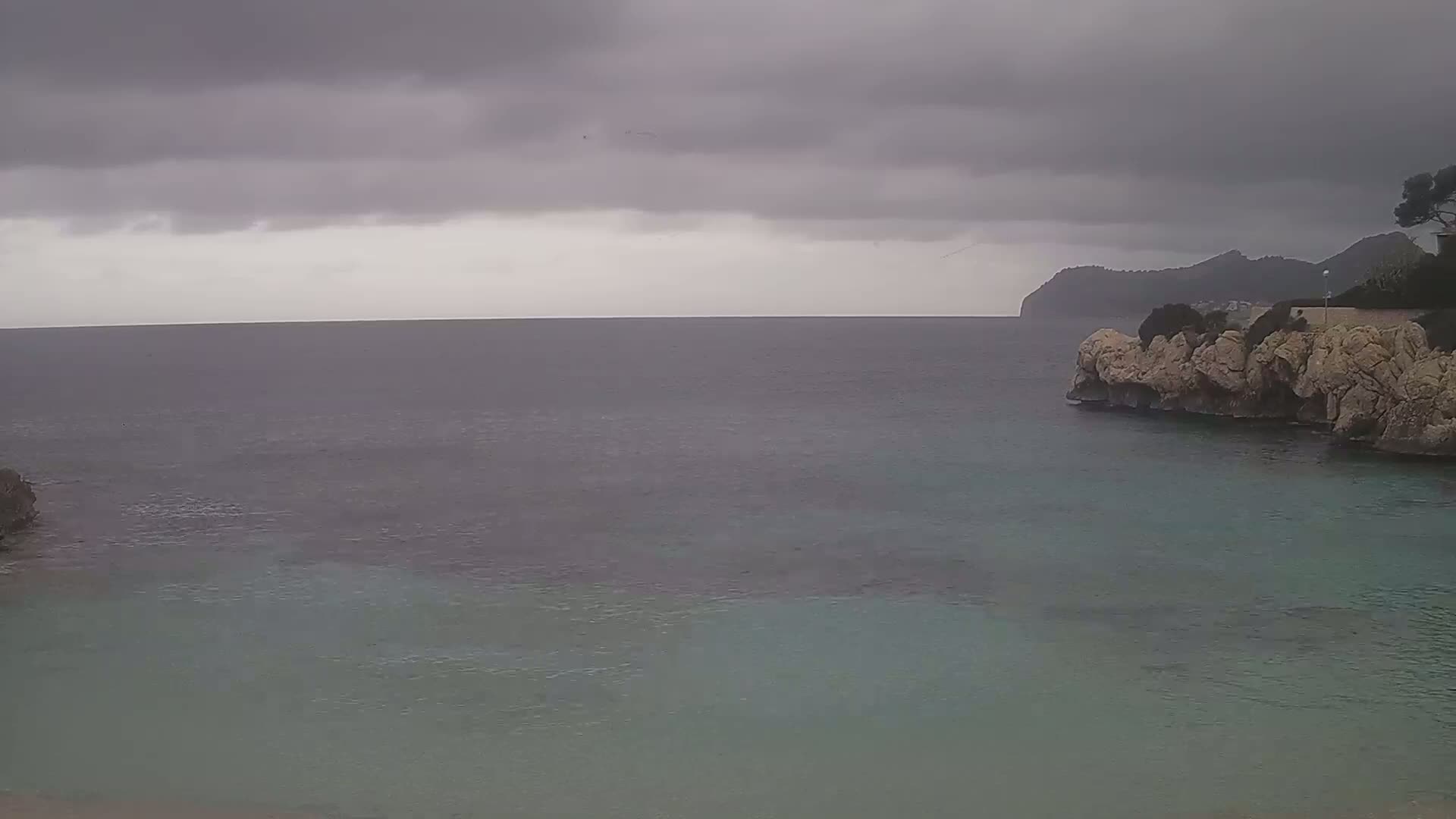 A calm sea, partially enclosed by rocky shores, is viewed under an overcast sky.