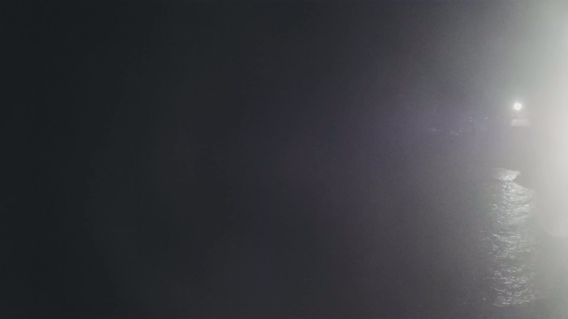 A dimly lit, foggy scene shows dark water reflecting a bright light in the distance.