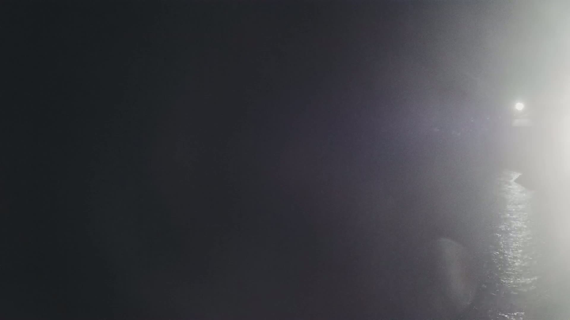 A dimly lit, foggy scene shows a dark body of water reflecting a light source in the distance.