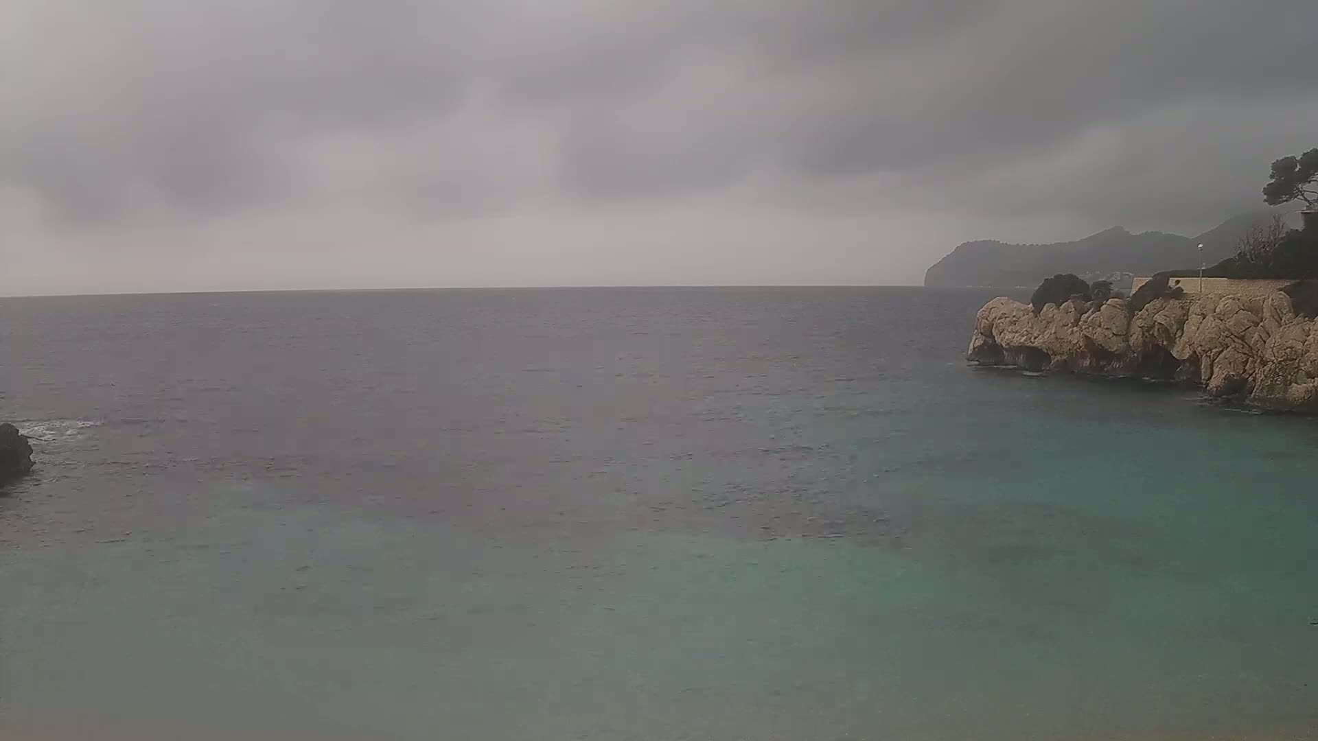A calm, teal sea laps against a rocky shore under an overcast sky, with a distant mountain visible in the background.