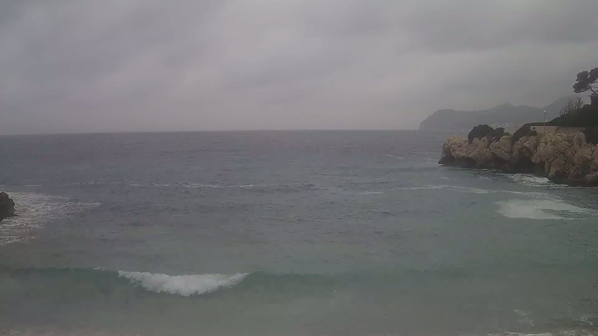 A gray, overcast day shows a rocky shoreline with small waves breaking on a calm sea, and distant hills under a cloudy sky.