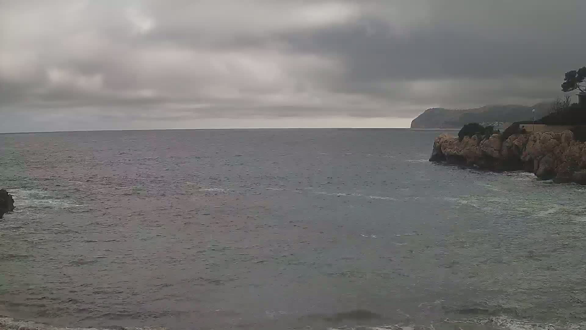 A gray, overcast sky hangs over a calm sea, partially enclosed by rocky shores, with a distant landmass visible on the horizon.