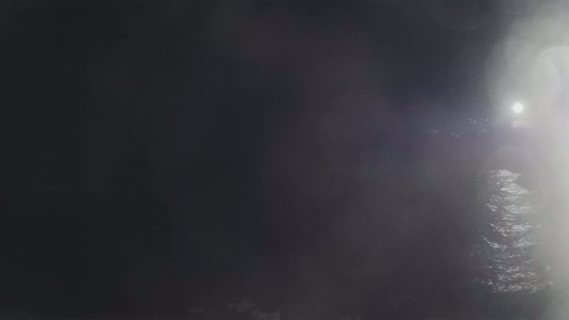 A blurry, nighttime image shows dark water with faint waves and a bright light in the distance, obscured by fog or mist.