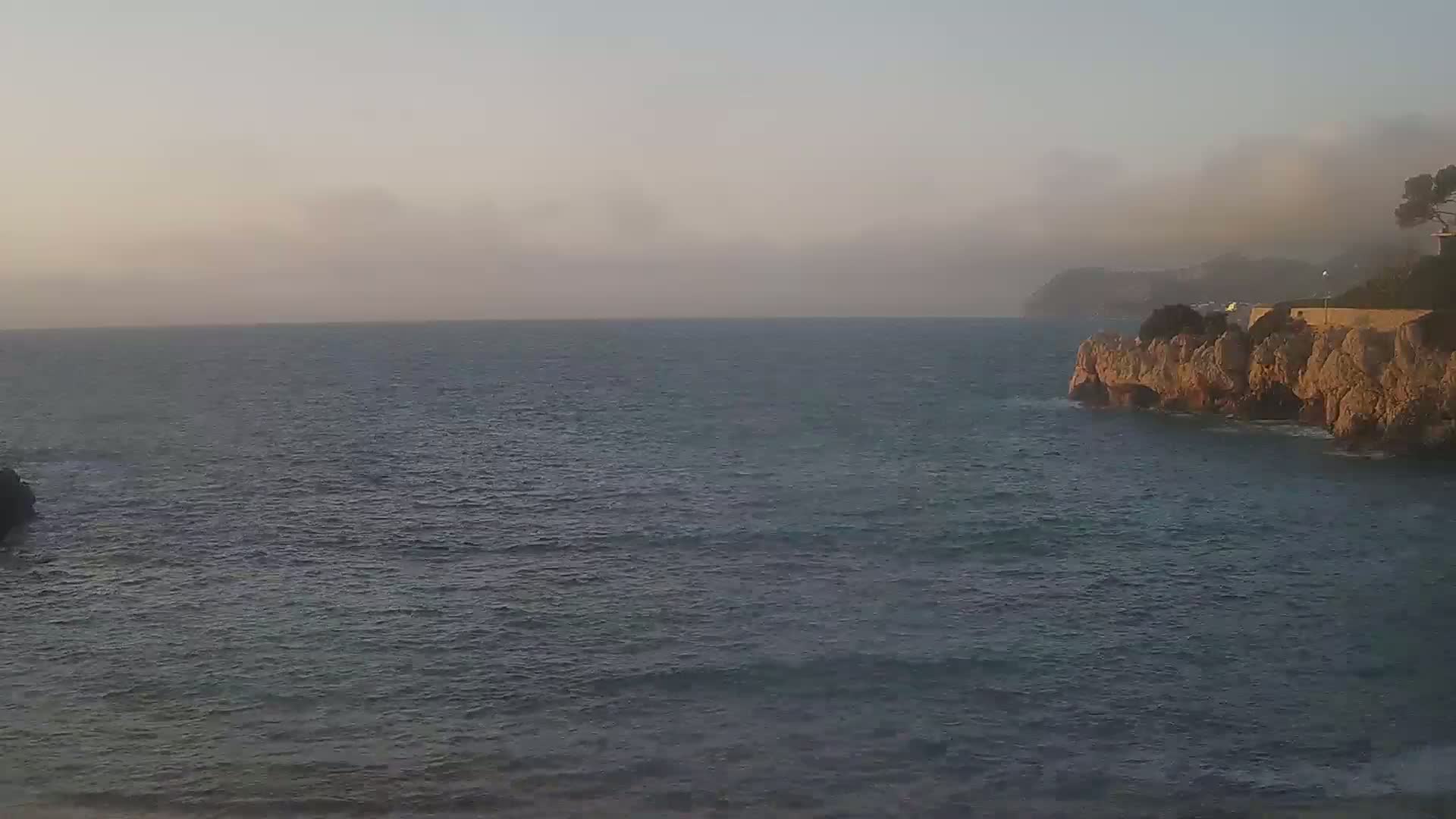 A calm sea meets a rocky cliff under a hazy, light sky.
