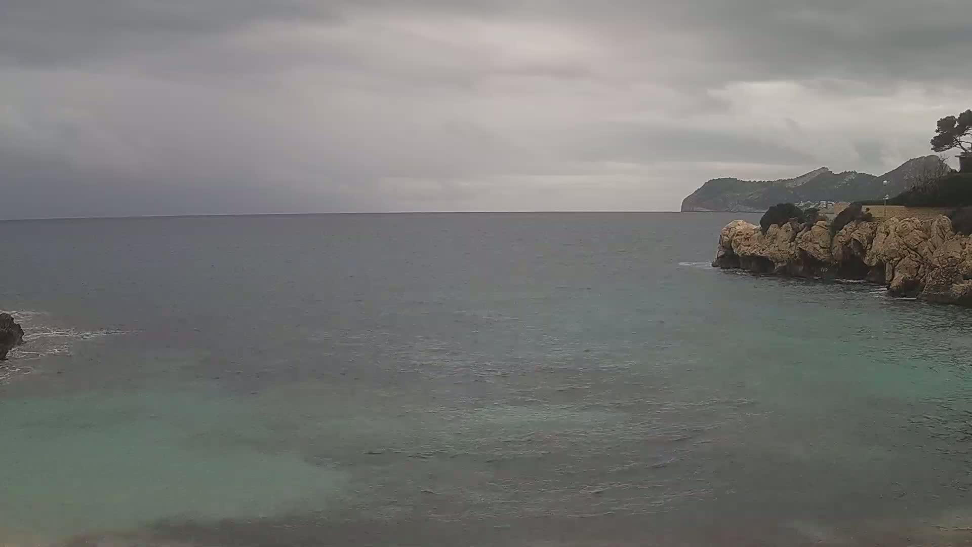 A calm, gray sea laps against rocky shores under an overcast sky, with a distant landmass visible in the background.