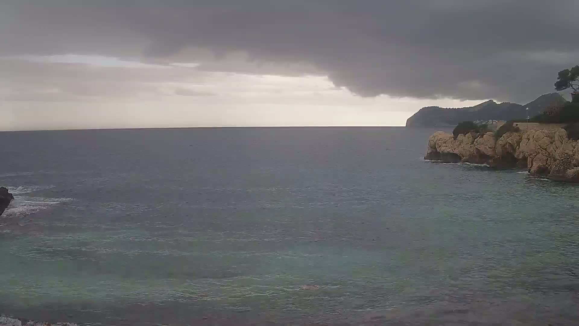A dark, overcast sky hangs over a calm sea meeting a rocky coastline with a distant hill visible.