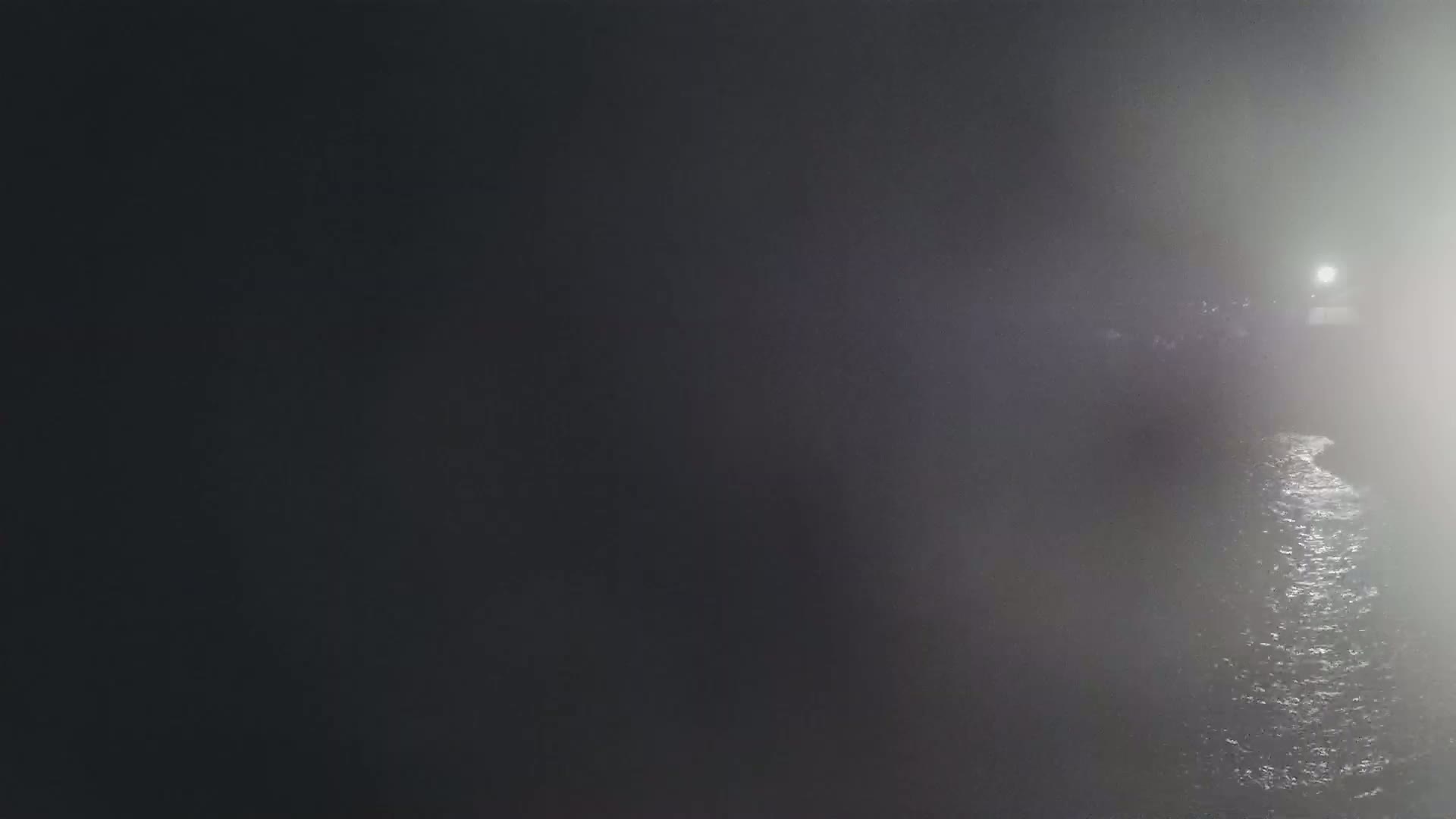 A dimly lit body of water is seen at night, shrouded in fog with a light source visible in the distance.
